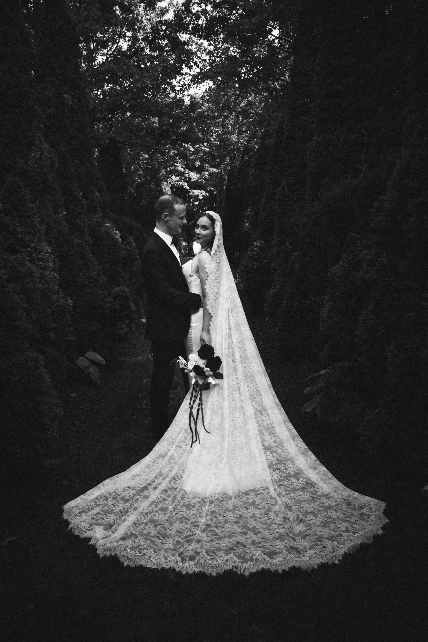 Black and white wedding photo of bride in lace gown with cathedral veil and groom in tuxedo embracing in lush garden.