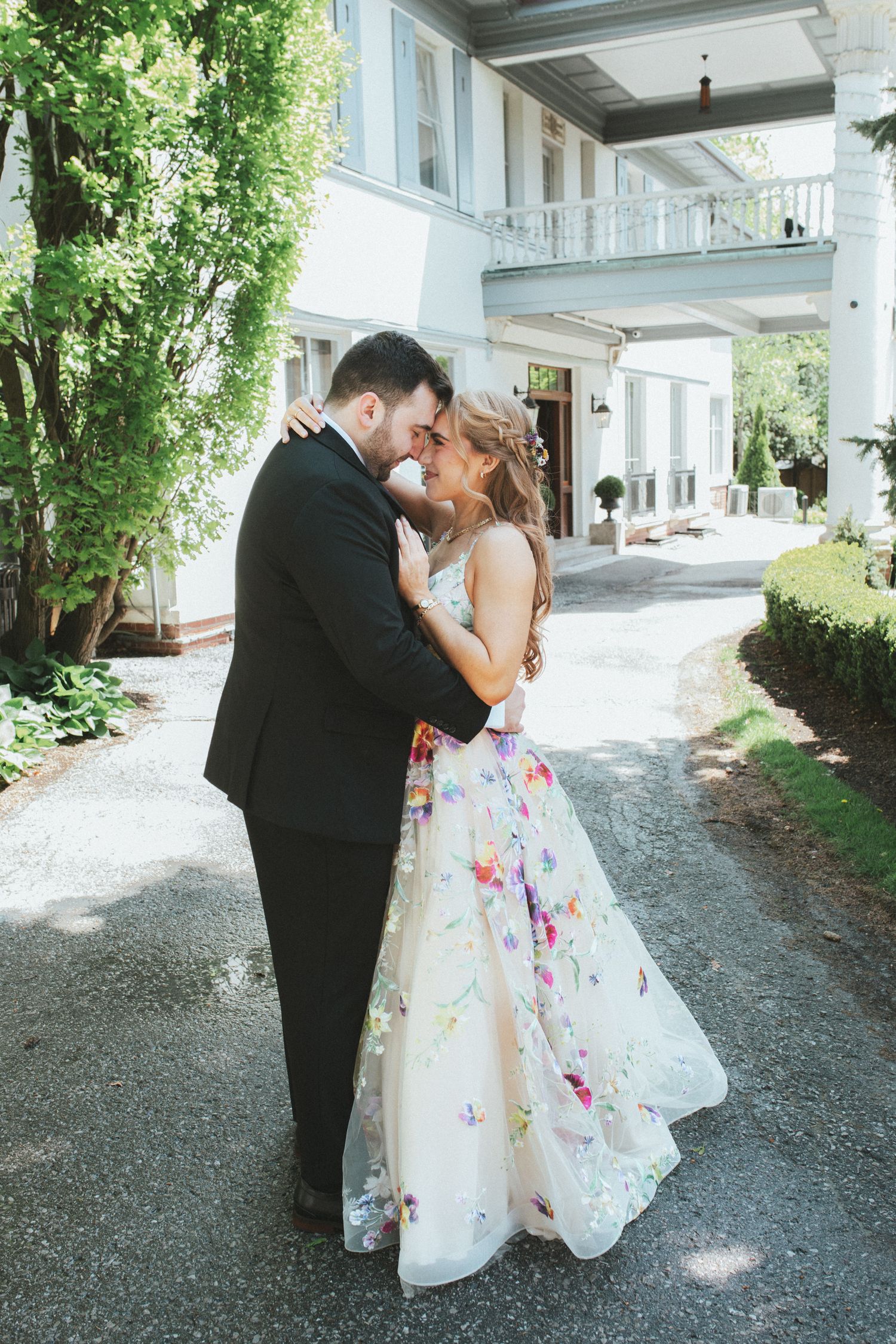 A couple shares an intimate moment on a tree-lined gravel path outside a white building.