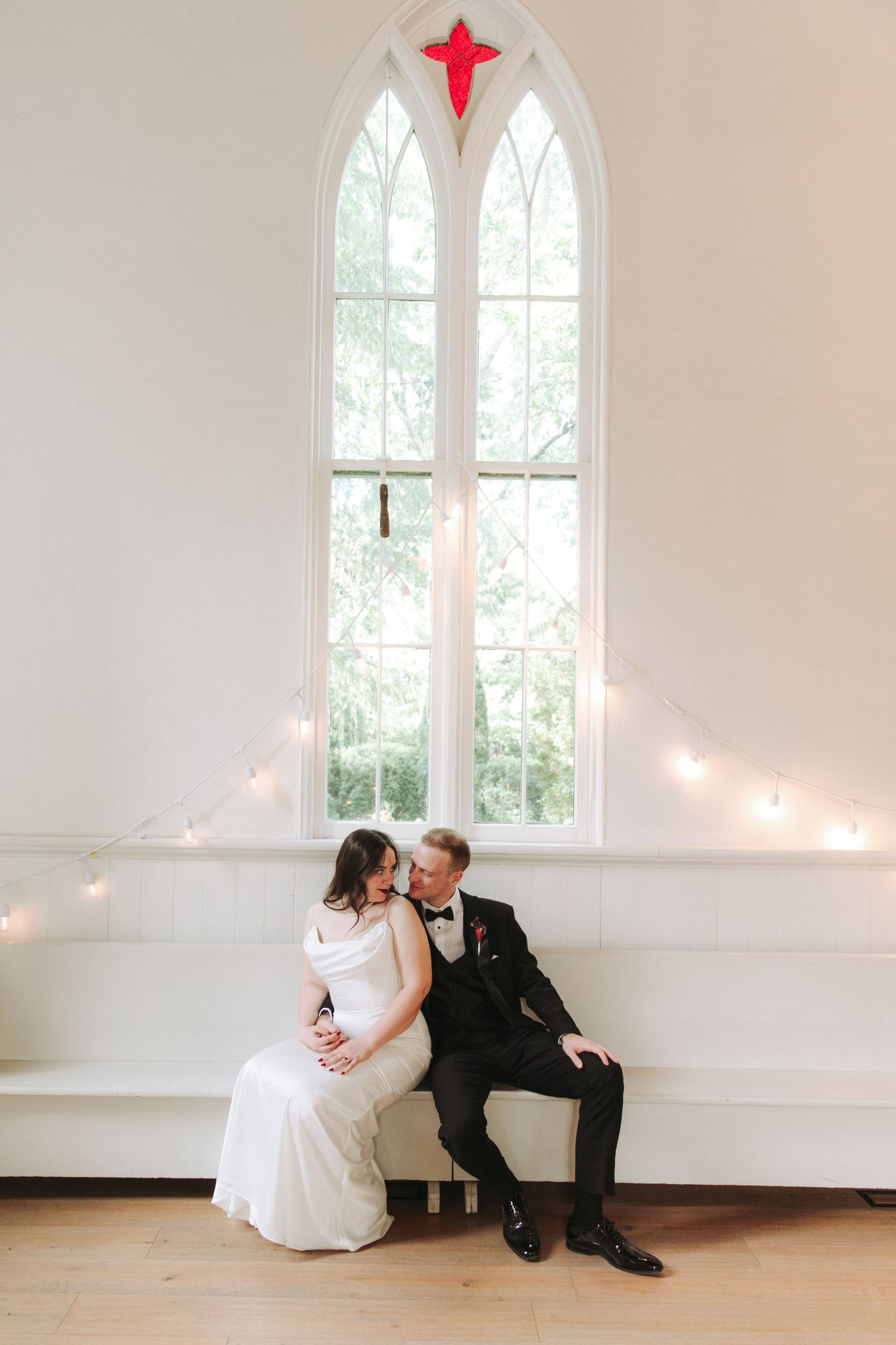 Bride and groom sitting together on a white bench beneath a tall arched window in a bright white chapel.