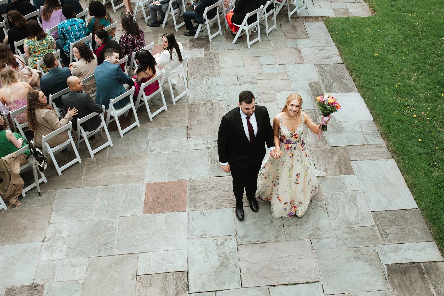 A couple walks down a stone path at an outdoor wedding ceremony while guests look on from white folding chairs.