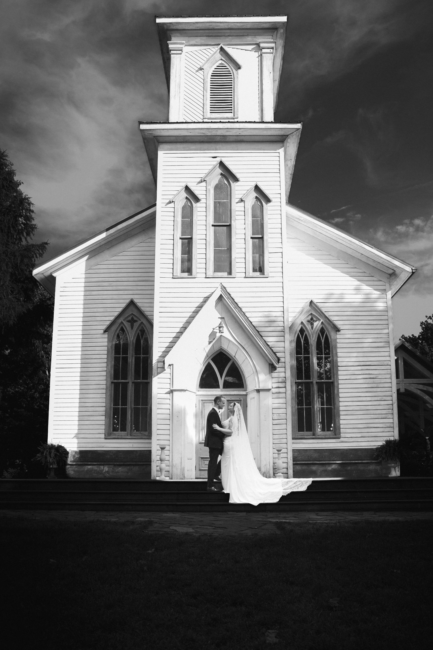 Black and white photo of a bride and groom embracing at the entrance of a classic white wooden church.
