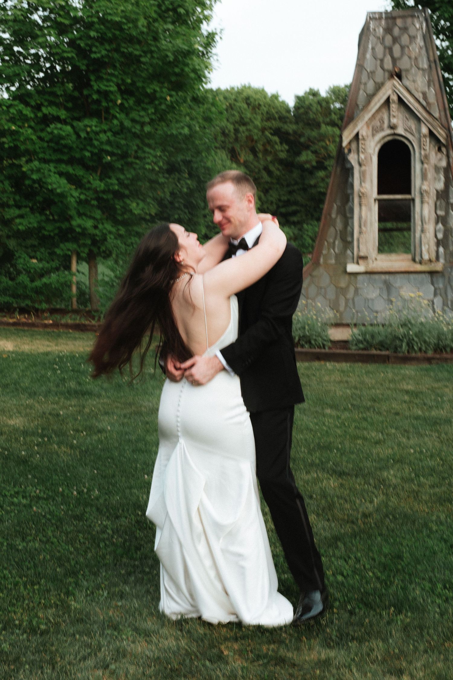 A bride and groom share a romantic dance outdoors on green grass near a stone chapel surrounded by lush trees.