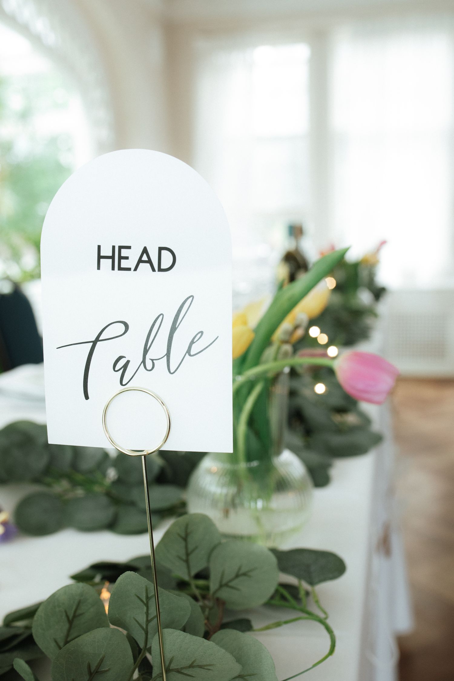 White head table sign with elegant calligraphy displayed in a holder among green foliage and floral centerpieces.