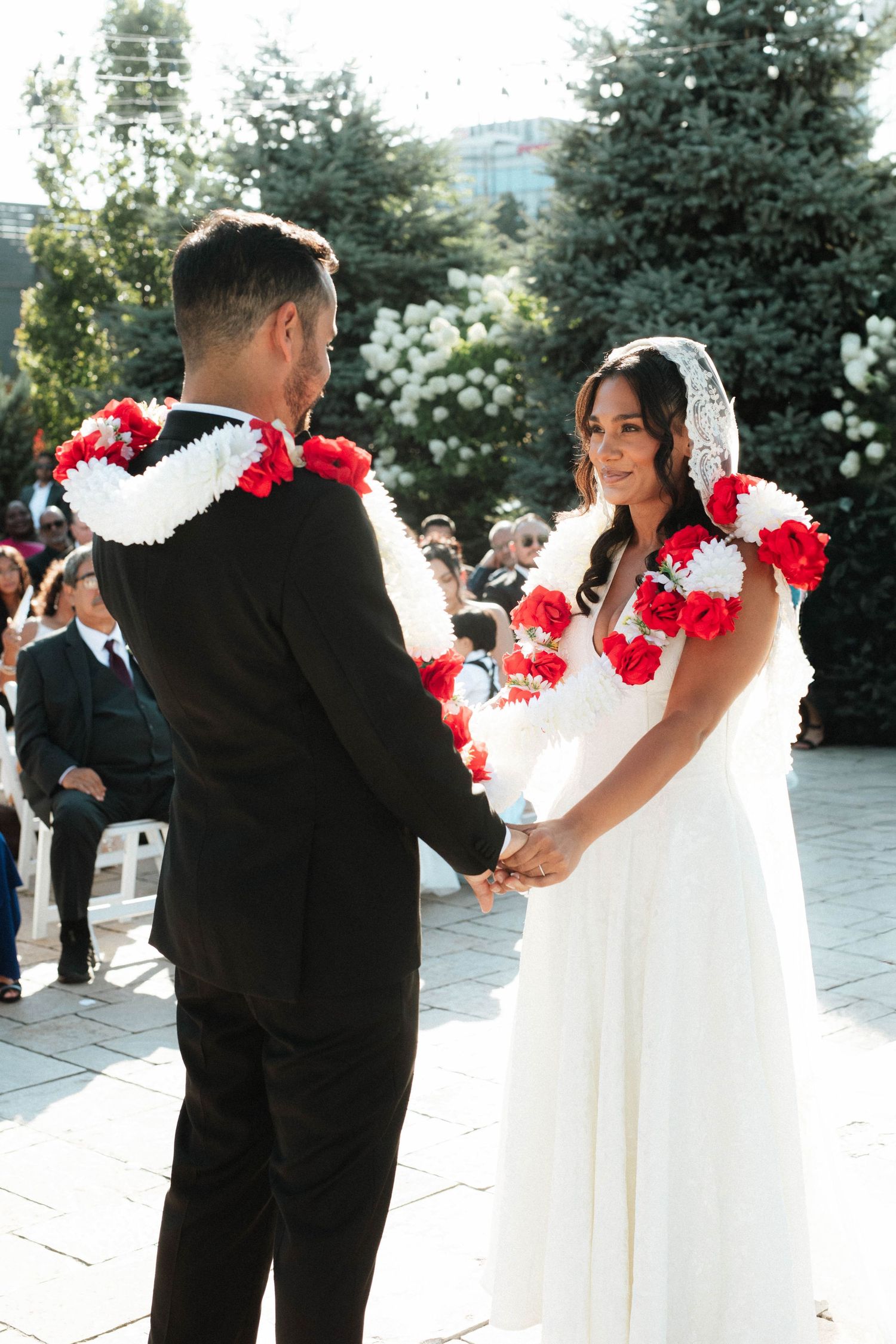 Bride and groom hold hands during outdoor wedding ceremony wearing red and white leis in bright sunlight.