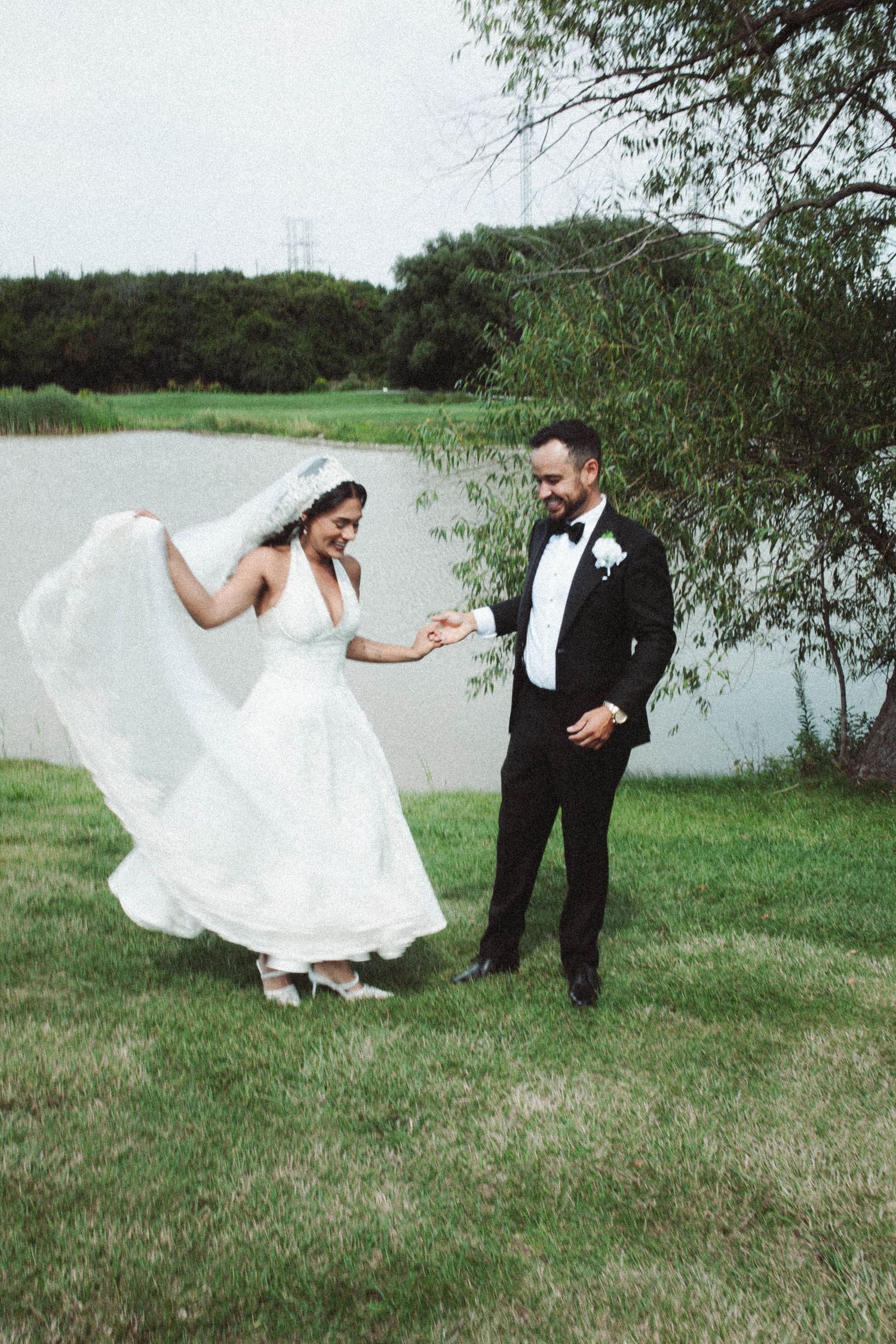 A bride's white dress flows in the wind as she dances with her groom by a lake on their wedding day.