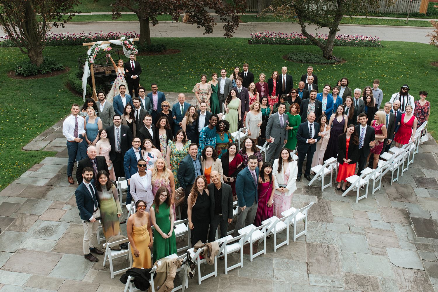 A panoramic view of wedding guests seated in white chairs on a stone patio surrounded by green lawn and trees.