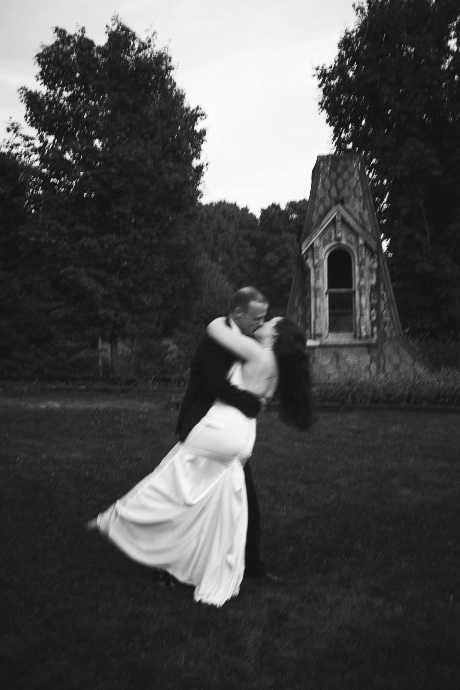 Black and white photo of a bride and groom sharing a romantic dip kiss outdoors near a historic stone chapel.
