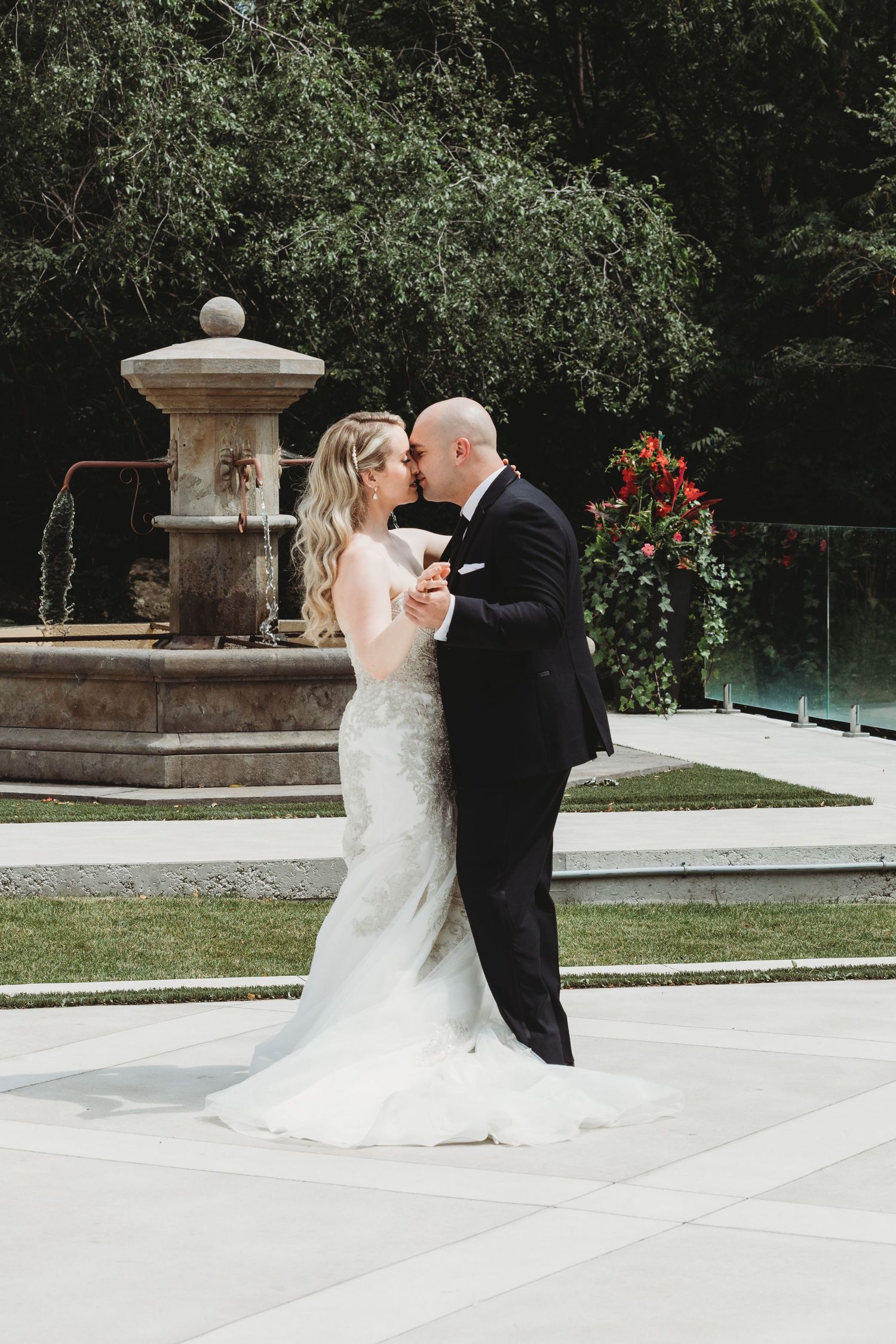 A romantic wedding couple shares a tender kiss outdoors on a stone patio surrounded by lush greenery and garden elements.