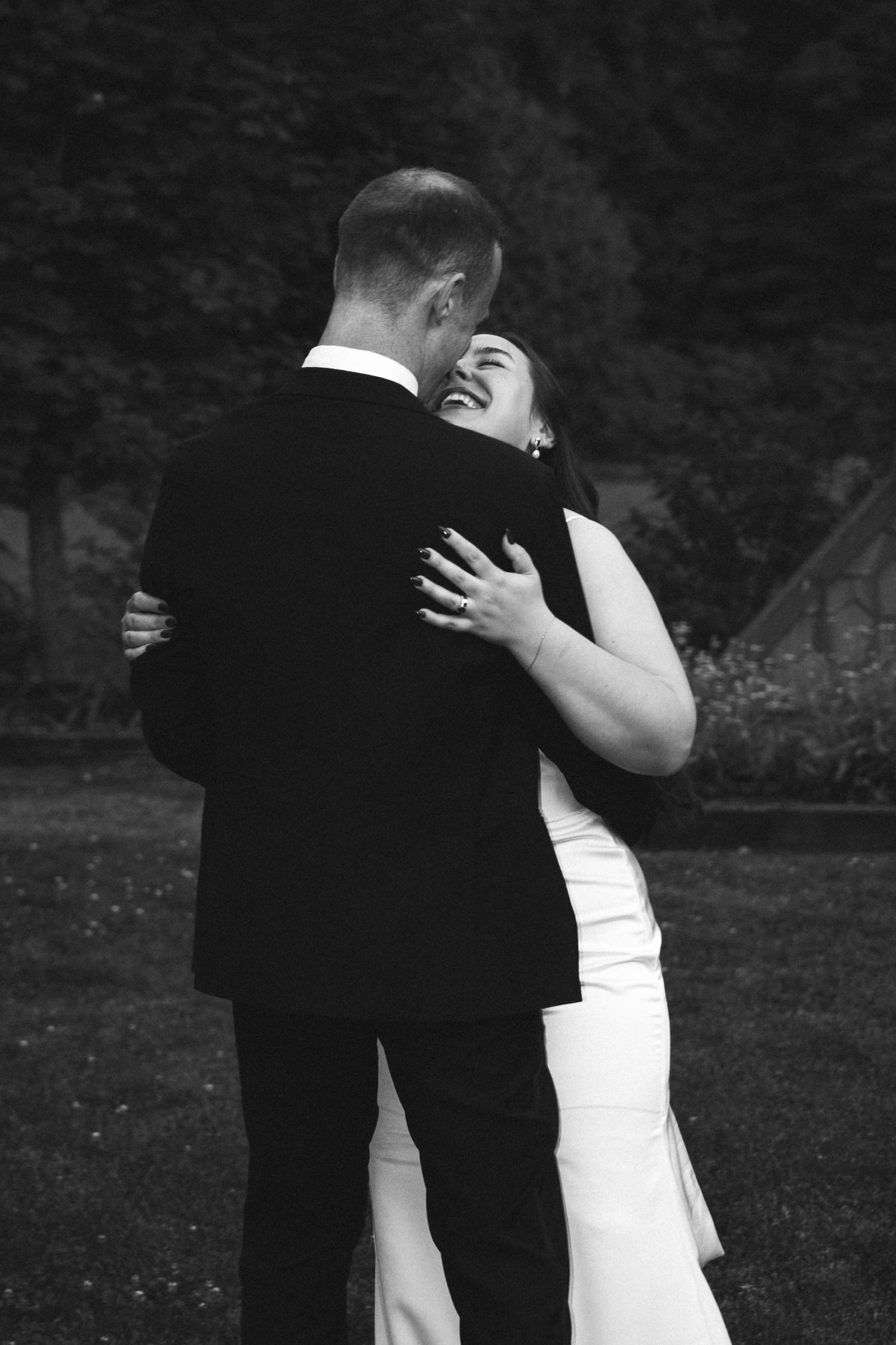 Black and white photo of a bride and groom embracing outdoors, the bride laughing joyfully in her wedding dress.