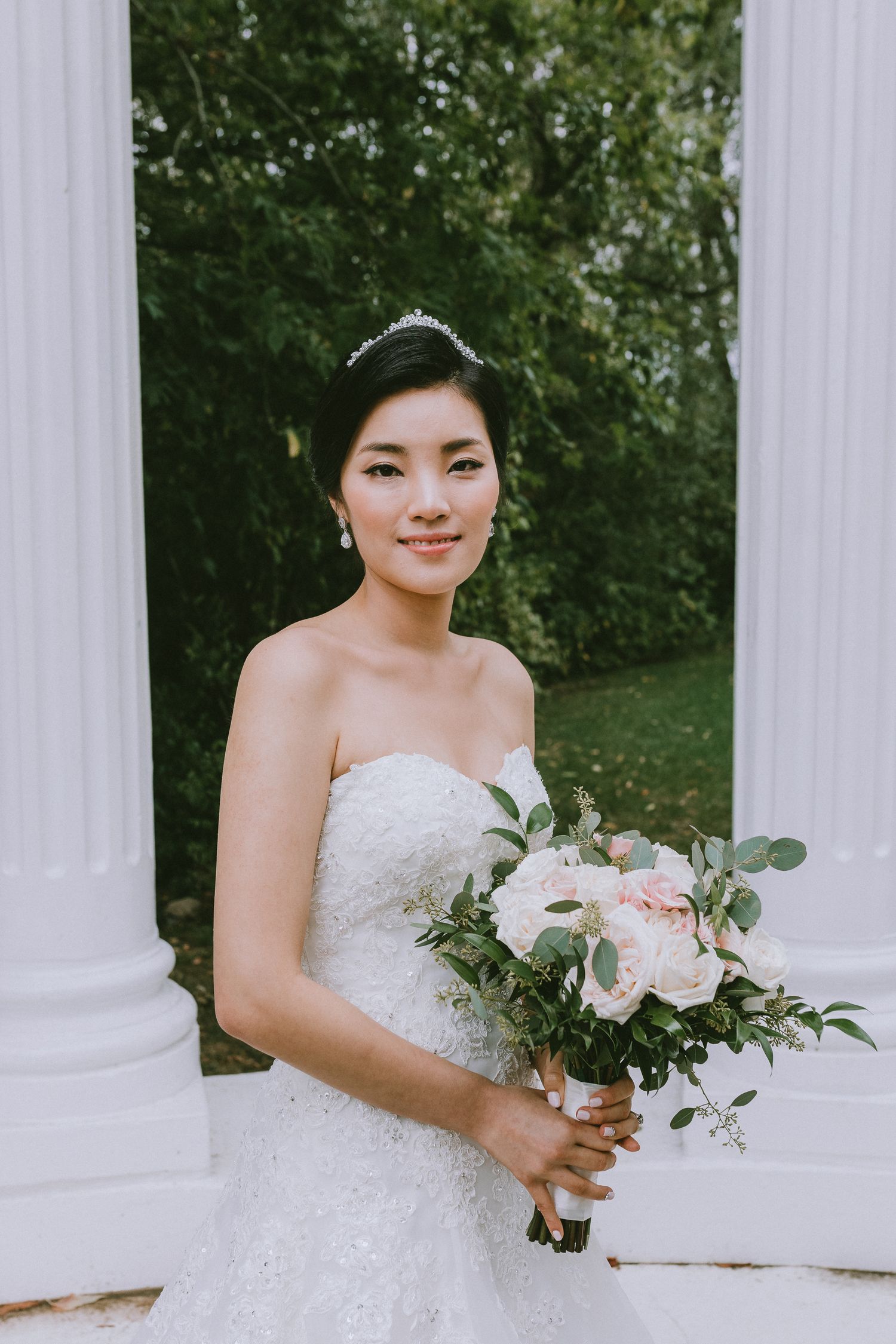 A bride in a white lace wedding dress holds a romantic pink and white floral bouquet against white columns.