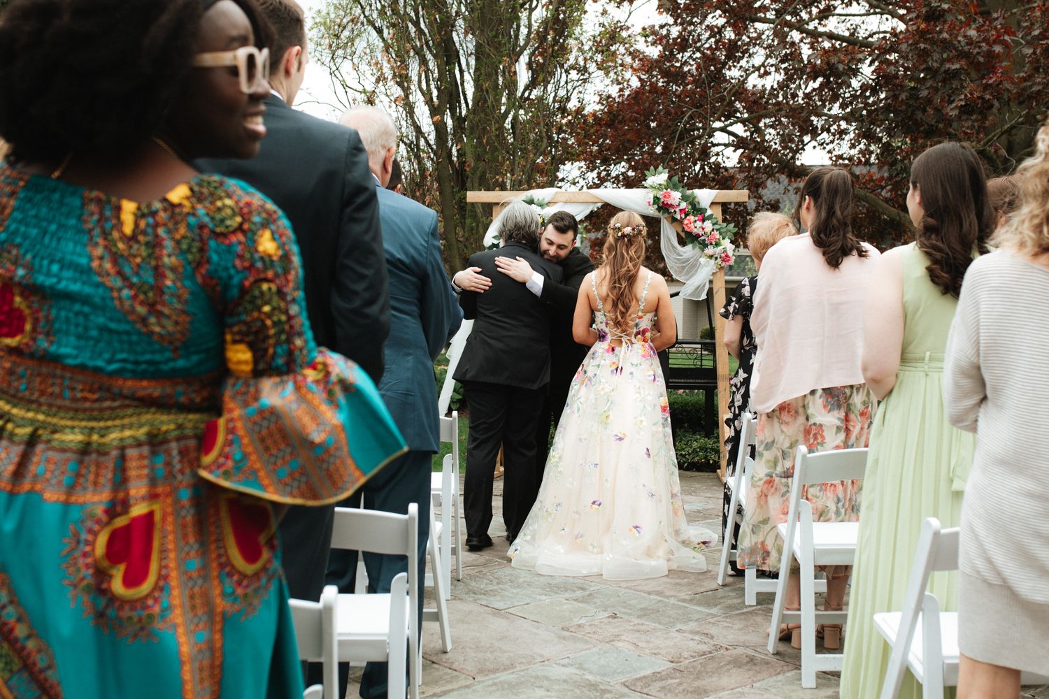 Wedding guests mingle outdoors during a garden ceremony with white chairs and decorative floral arrangements.