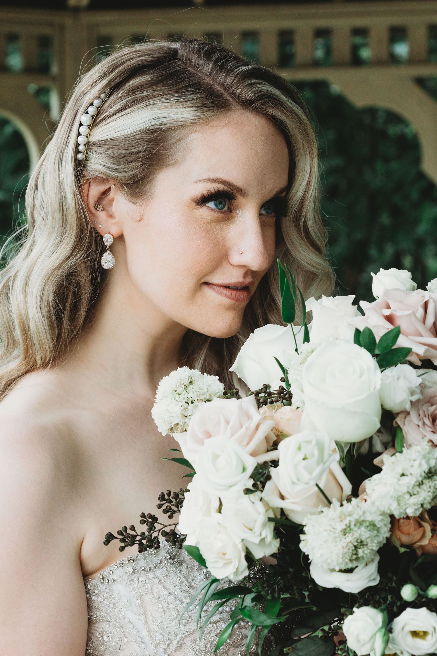 Close-up portrait of a bride with wavy blonde hair wearing a pearl headband and holding a bouquet of white roses.