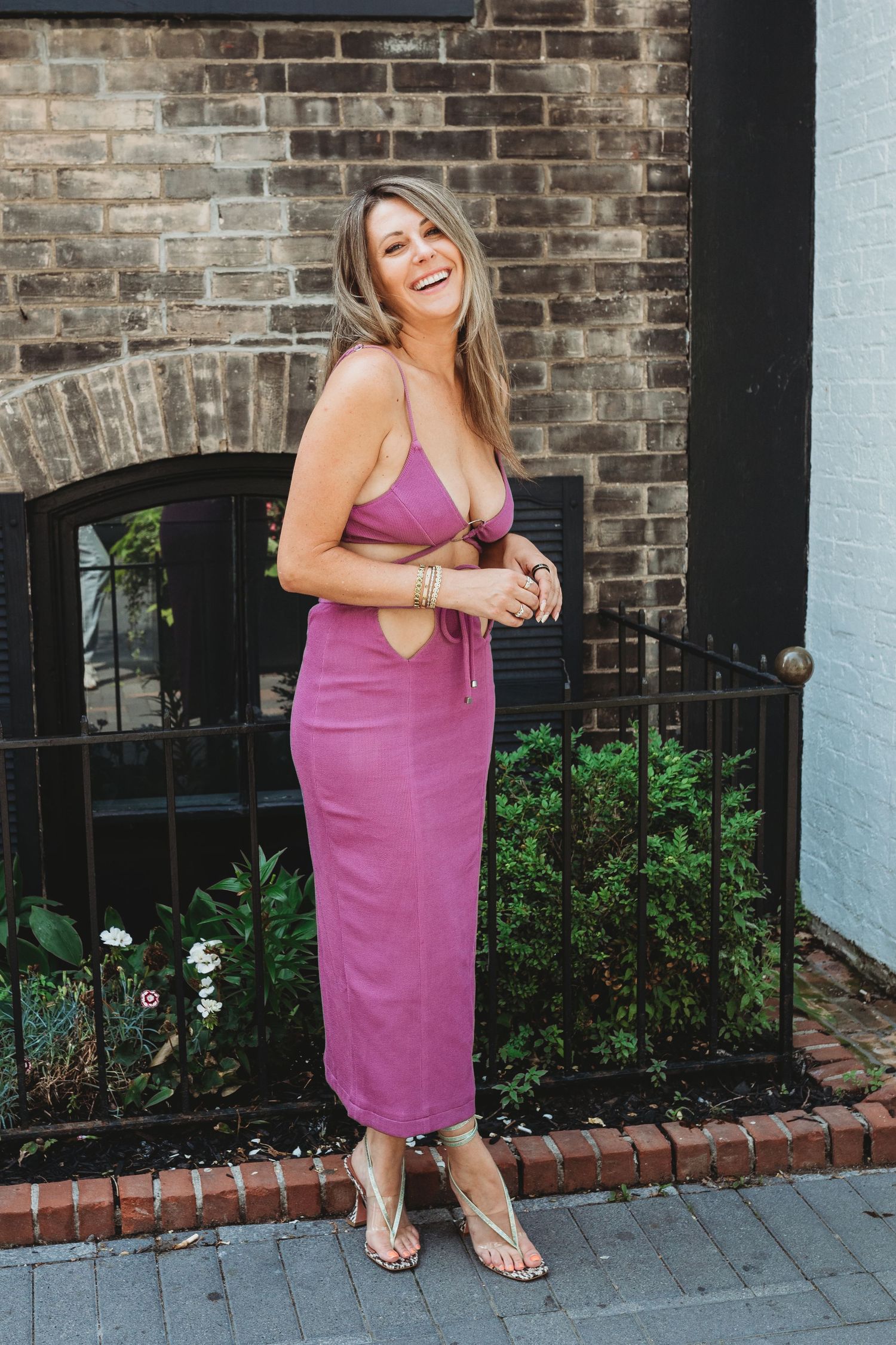 Woman in pink dress laughs joyfully while standing against brick wall with garden landscaping.