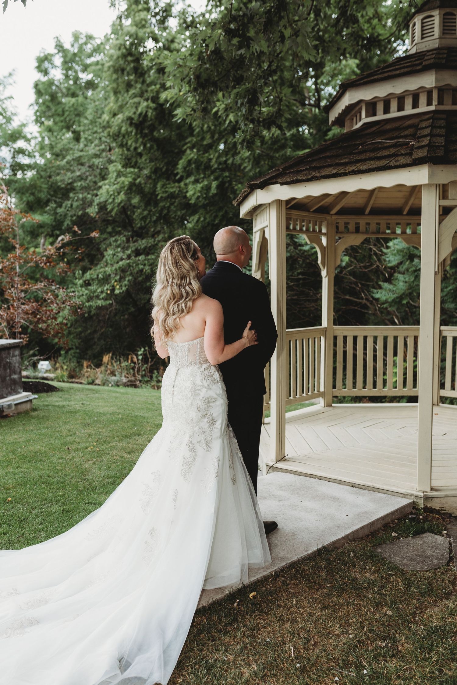 A couple in wedding attire share an intimate moment on a gazebo porch surrounded by lush green gardens.
