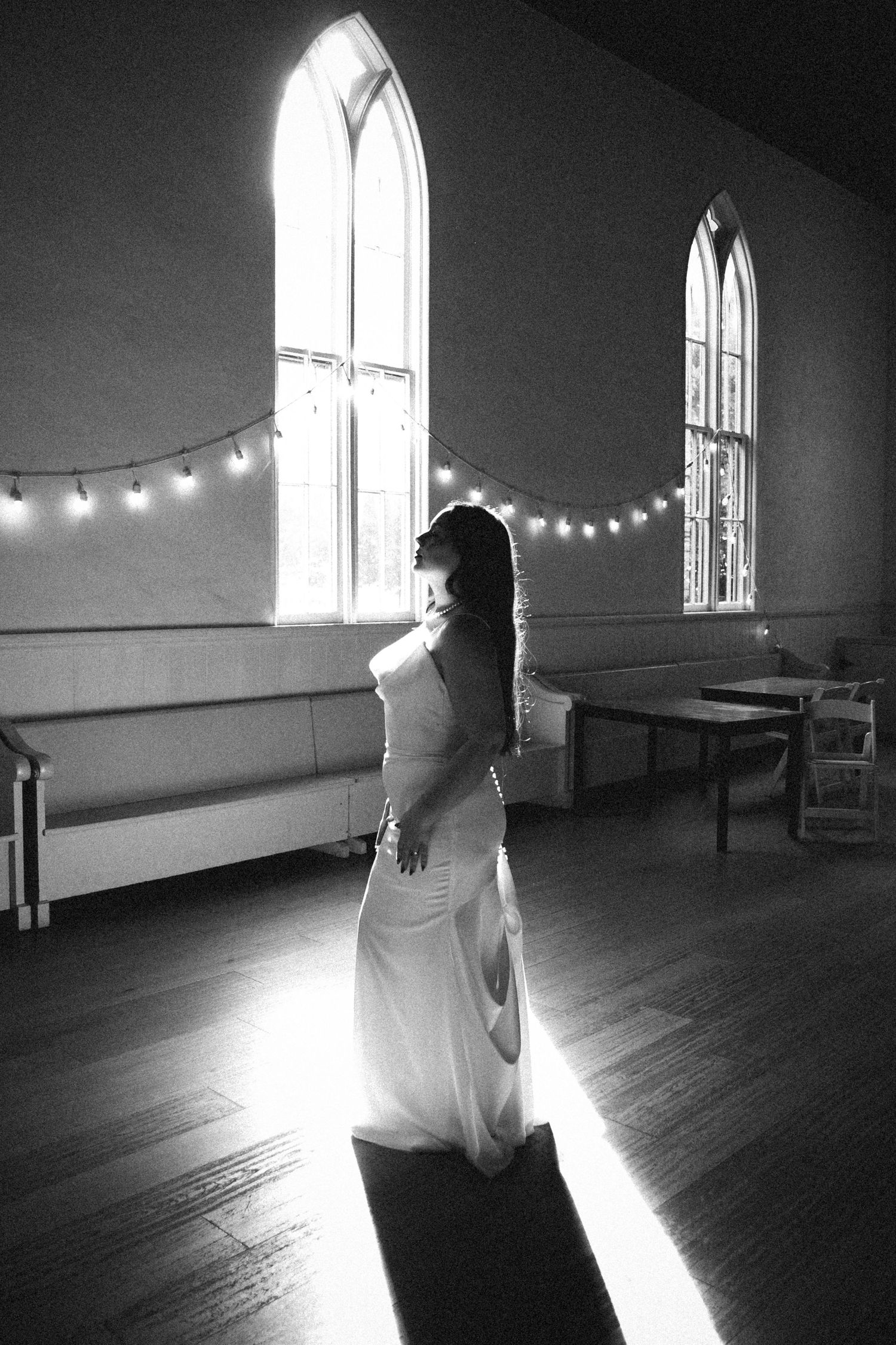 Bride in white gown standing in sunlit church hall with arched windows and string lights, black and white photo.