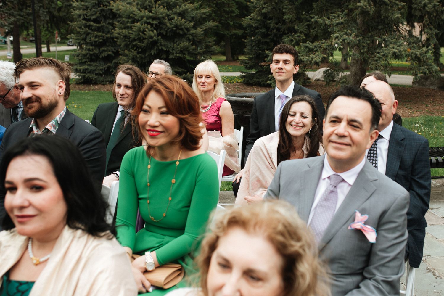 Wedding guests seated outdoors at a garden ceremony smile and look ahead during a sunny celebration.