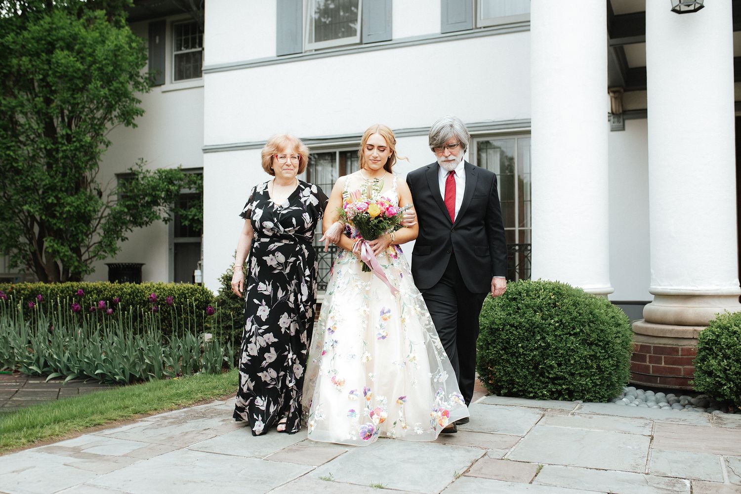 Wedding party poses outside white building entrance during outdoor summer celebration.