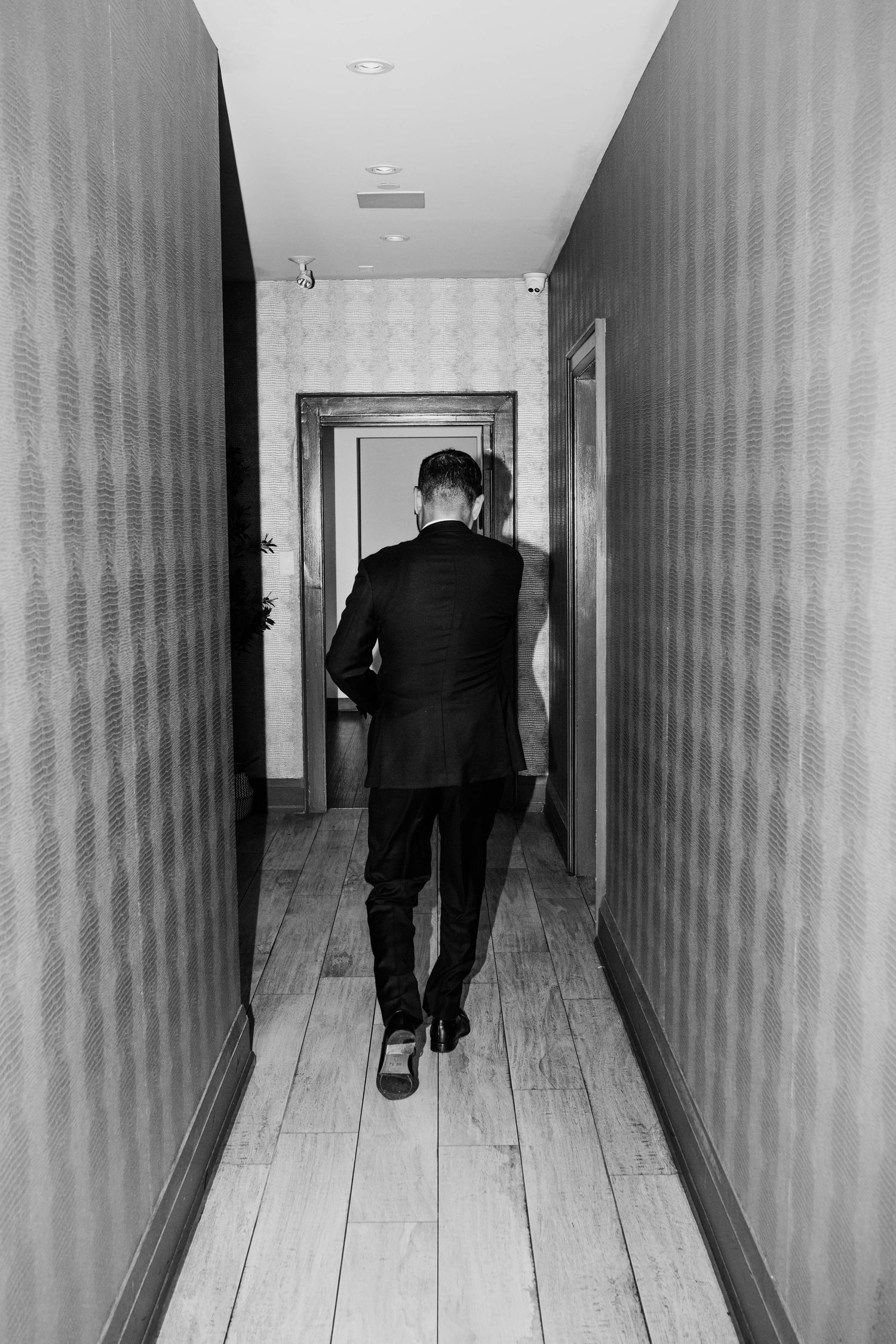 Black and white photo of a person in a suit walking down a narrow hallway with wooden floors and textured walls.