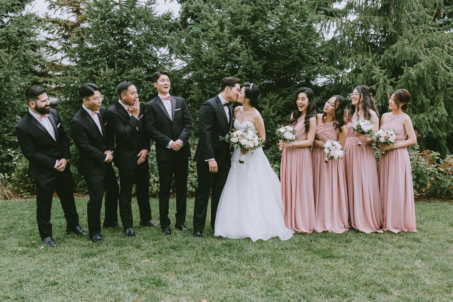 Wedding party poses outdoors with bridesmaids in pink dresses and groomsmen in black suits on a grassy lawn.