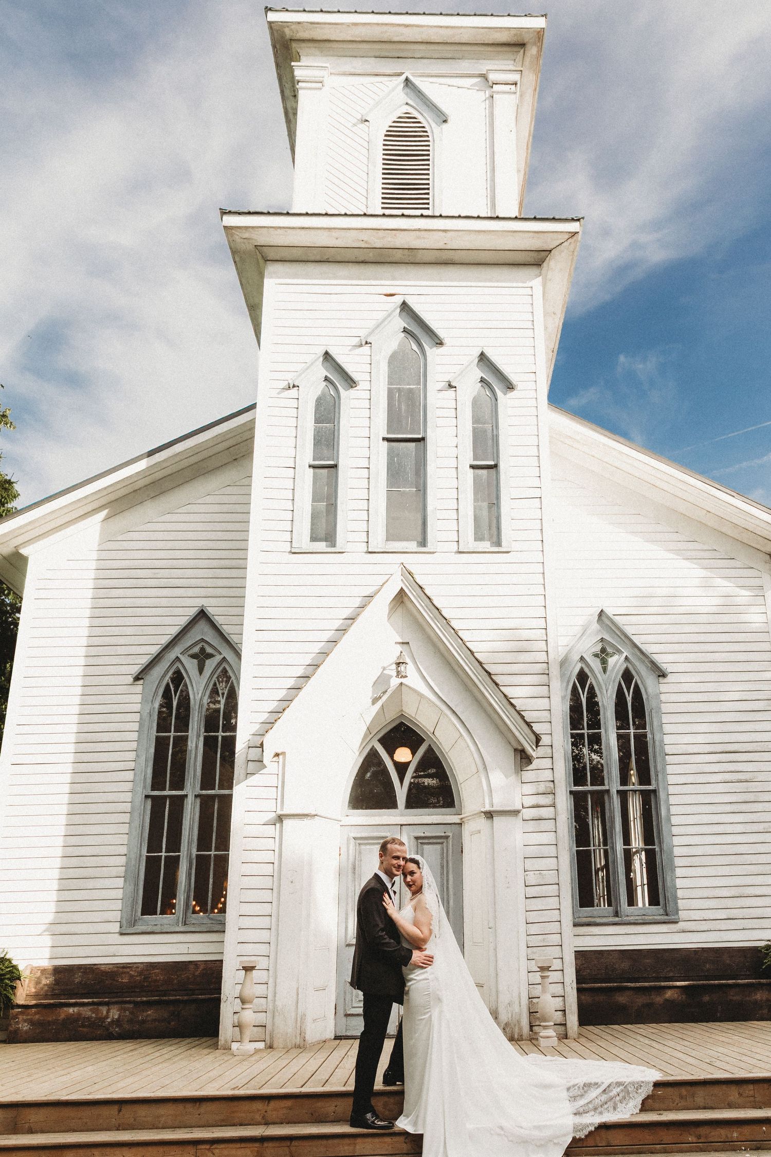 Bride and groom embracing in front of a classic white wooden church with Gothic arched windows on their wedding day.