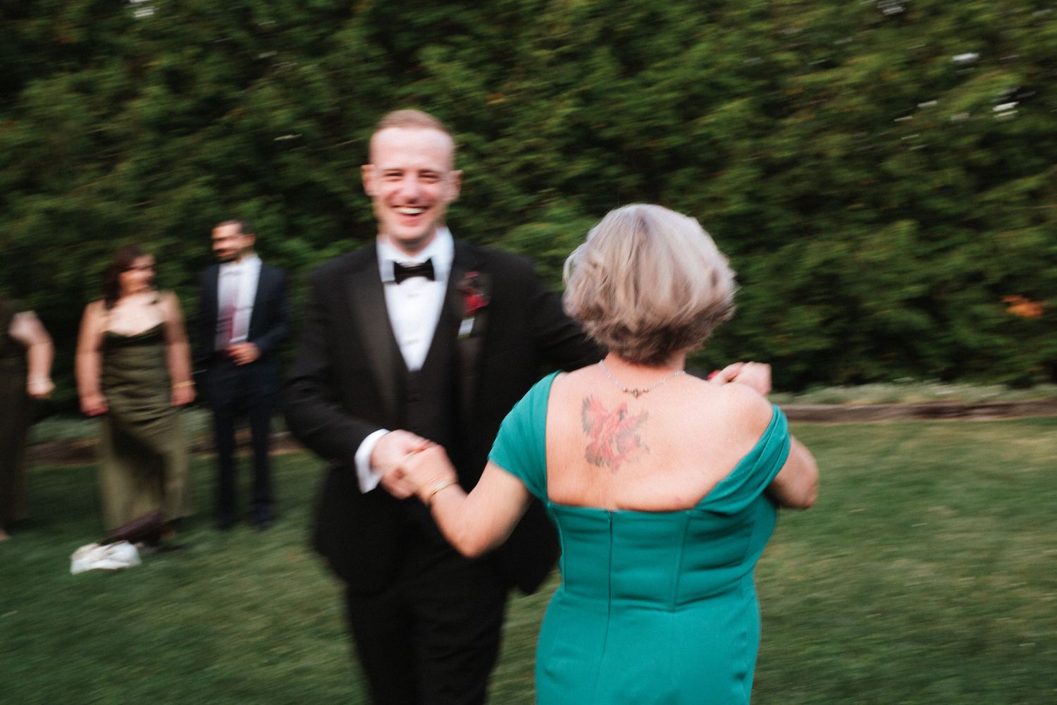Groom in tuxedo dancing with woman in teal dress and floral tattoo at outdoor wedding reception.