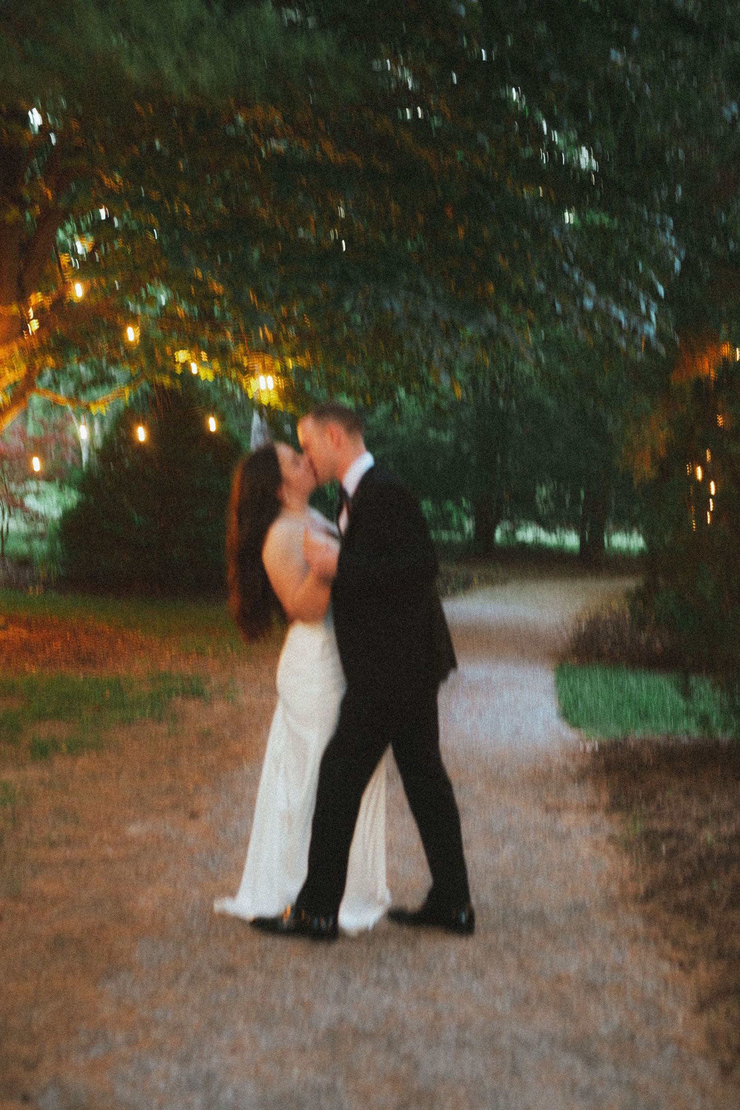 A bride and groom share a romantic kiss on a garden path at dusk, surrounded by glowing string lights and lush trees.