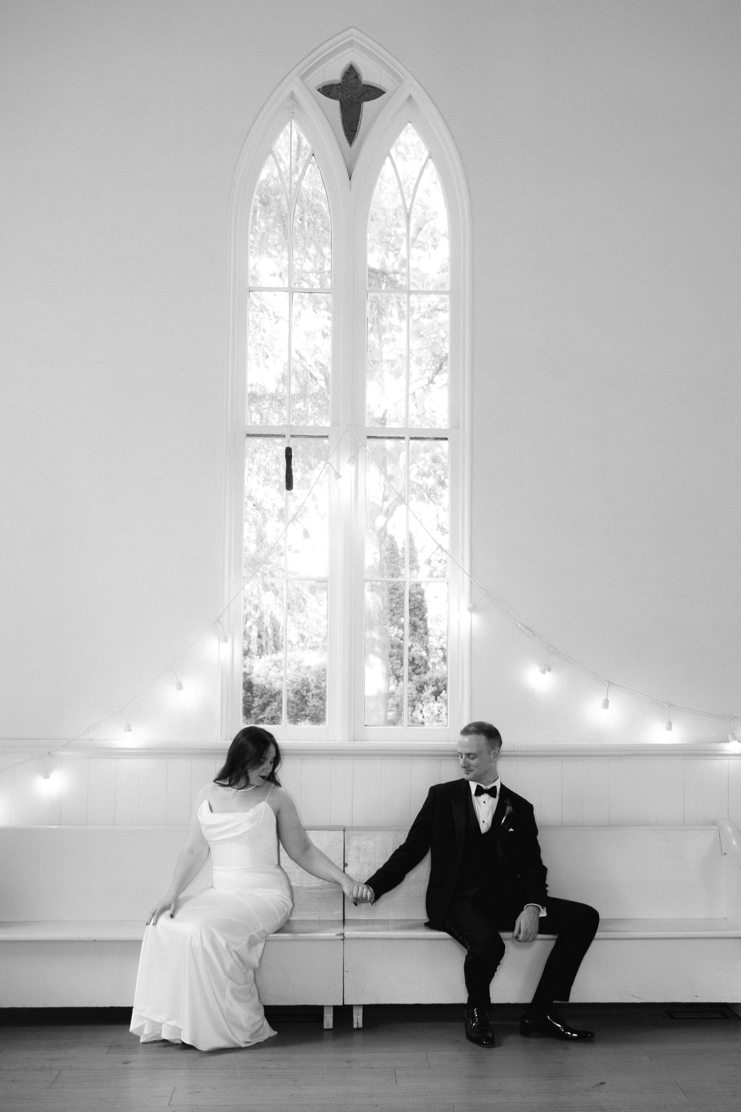 Black and white wedding photo of bride and groom holding hands on a bench beneath a gothic arch window in a white chapel.