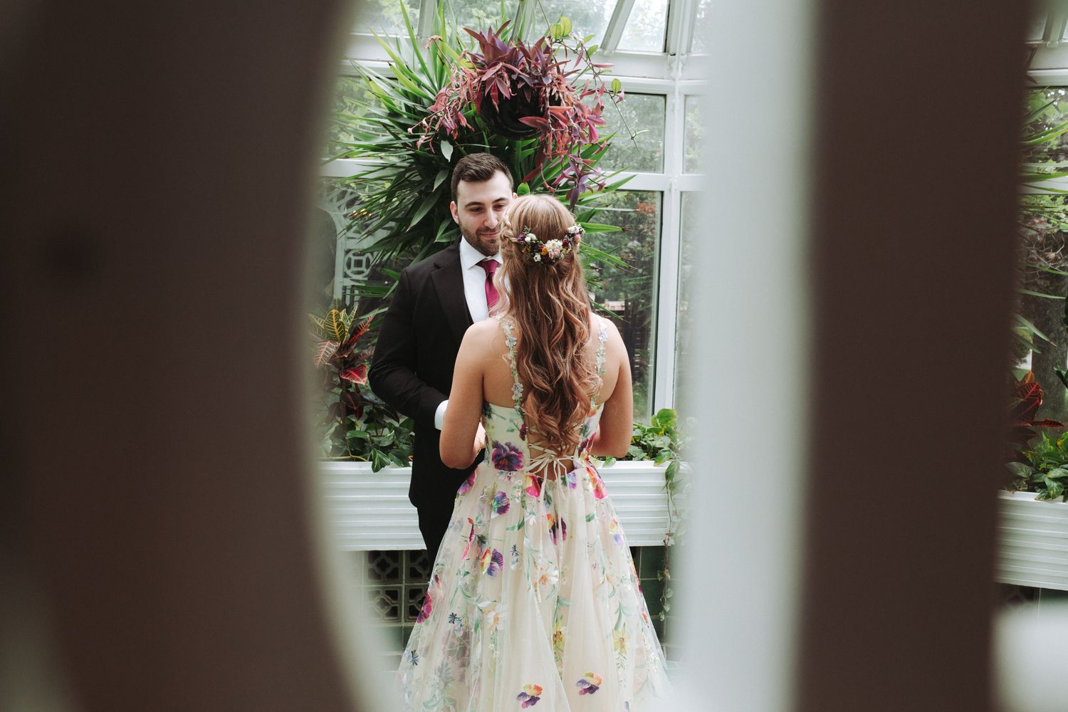 A bride and groom share an intimate moment in front of a window with greenery and pink floral decorations.