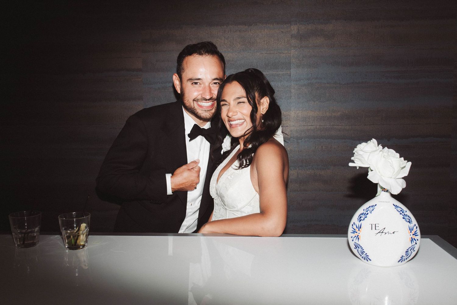 A happy couple in formal wedding attire embrace and smile while celebrating at their reception.
