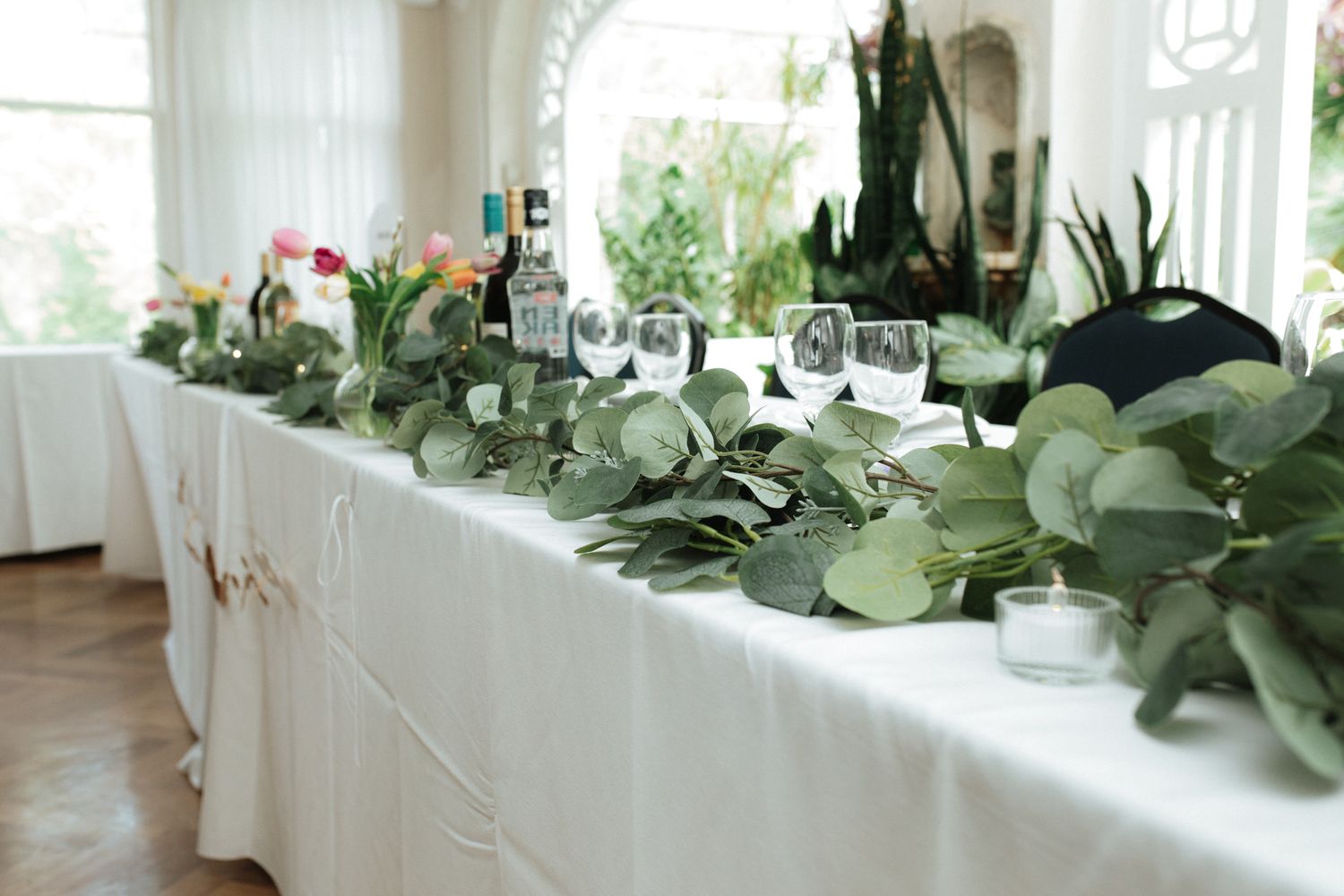 Elegant wedding reception table decorated with white linens and fresh eucalyptus greenery garland in natural light.