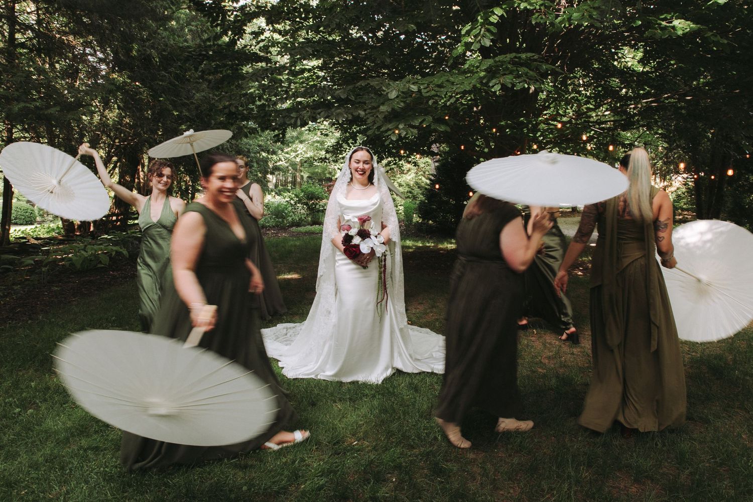 Bride in white gown with bridesmaids in green dresses spinning parasols outdoors in a lush garden wedding celebration.