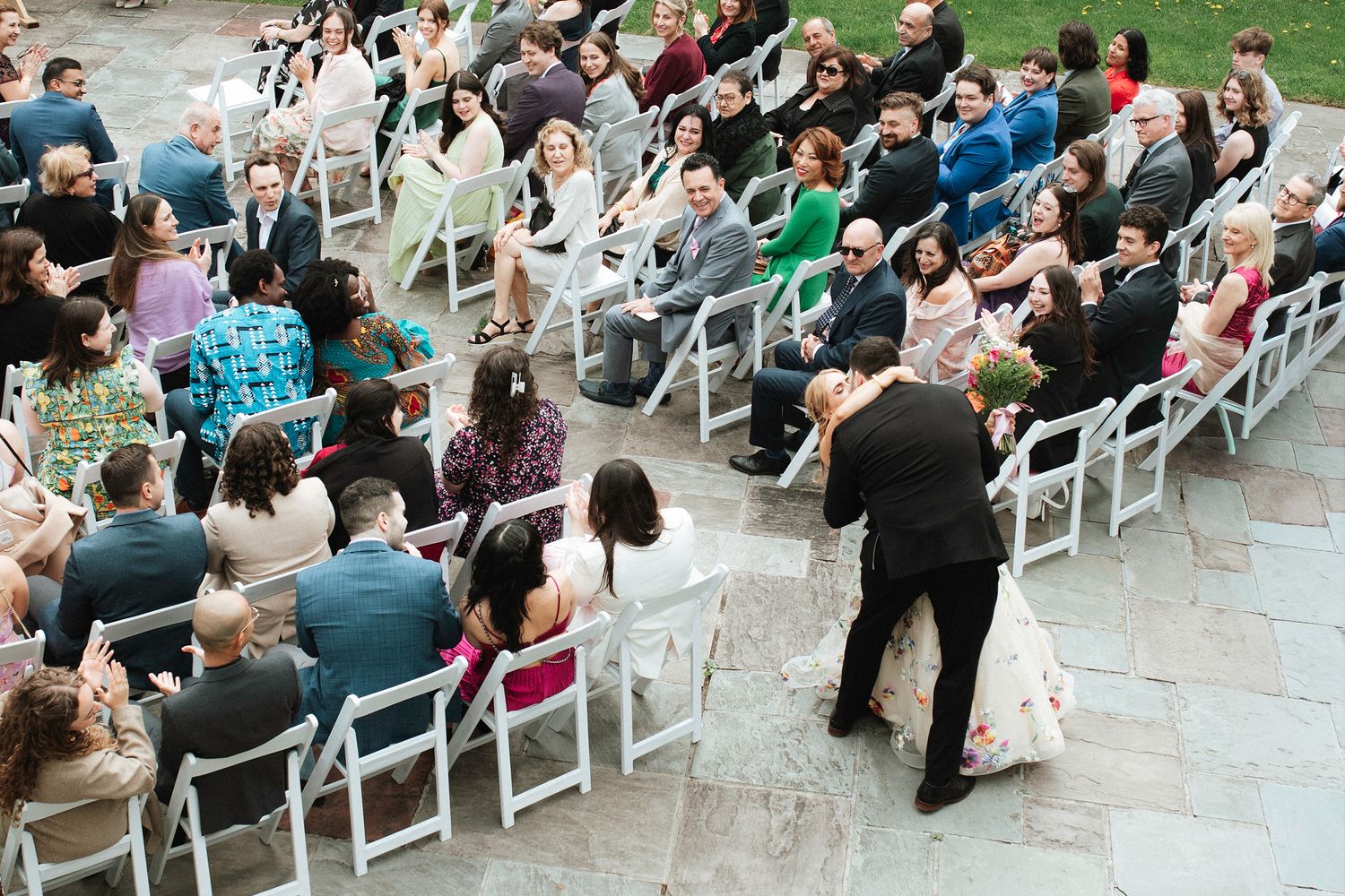 Guests seated in white chairs at an outdoor wedding ceremony on a stone patio.