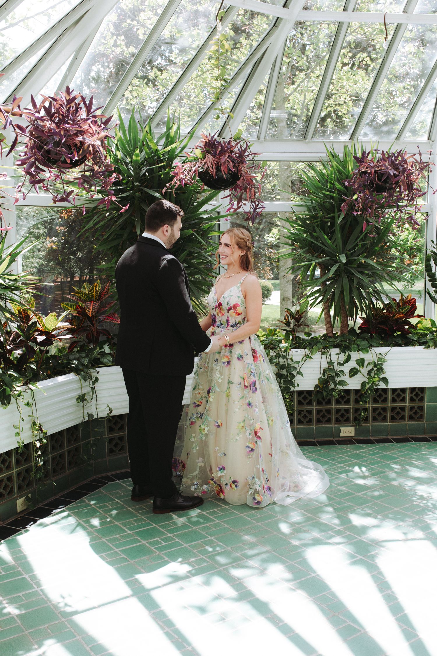 A romantic first look moment captured in a sunlit greenhouse with tropical plants and white lattice walls.