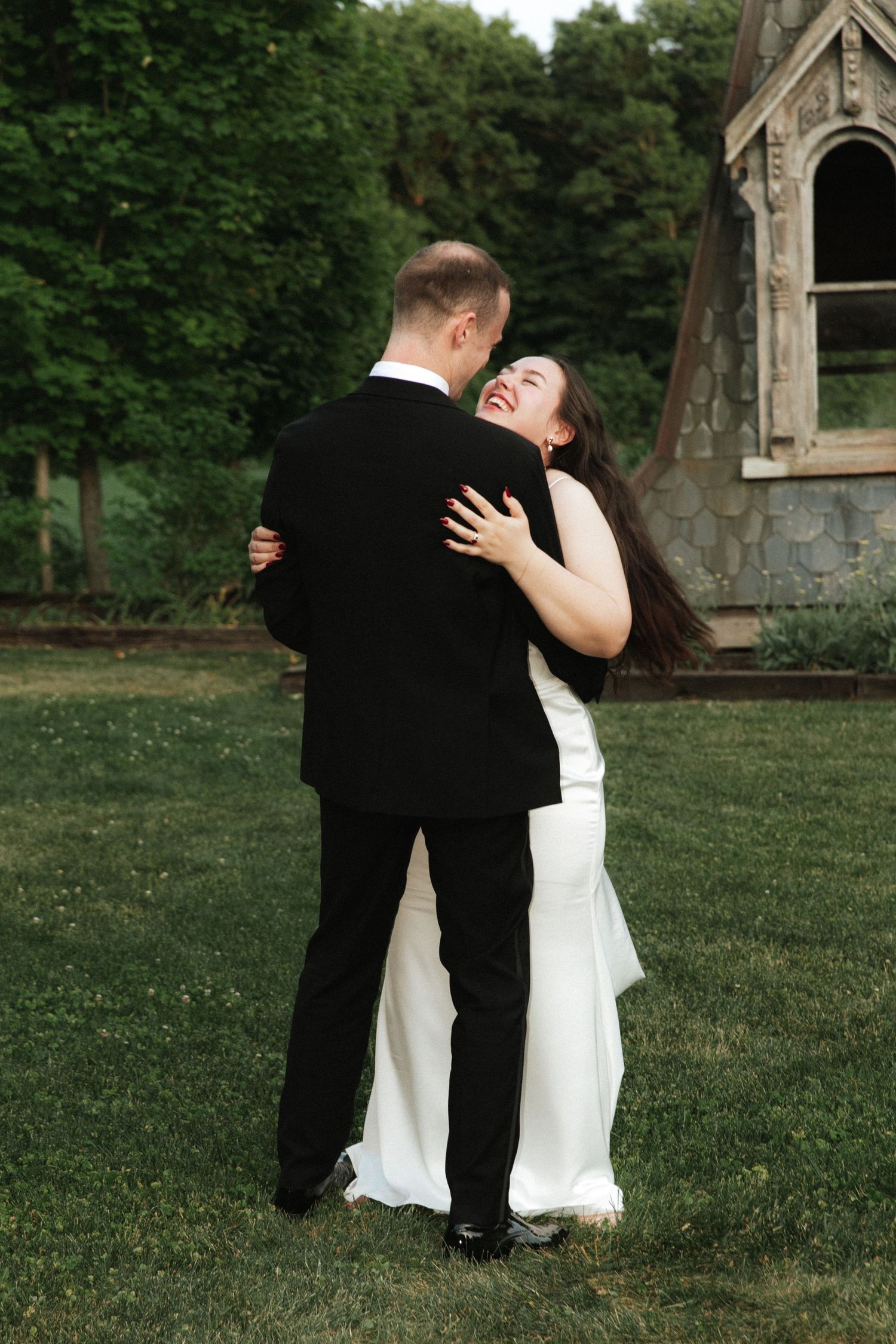 Bride in white gown laughing joyfully while embracing groom in black tuxedo outdoors on green lawn.