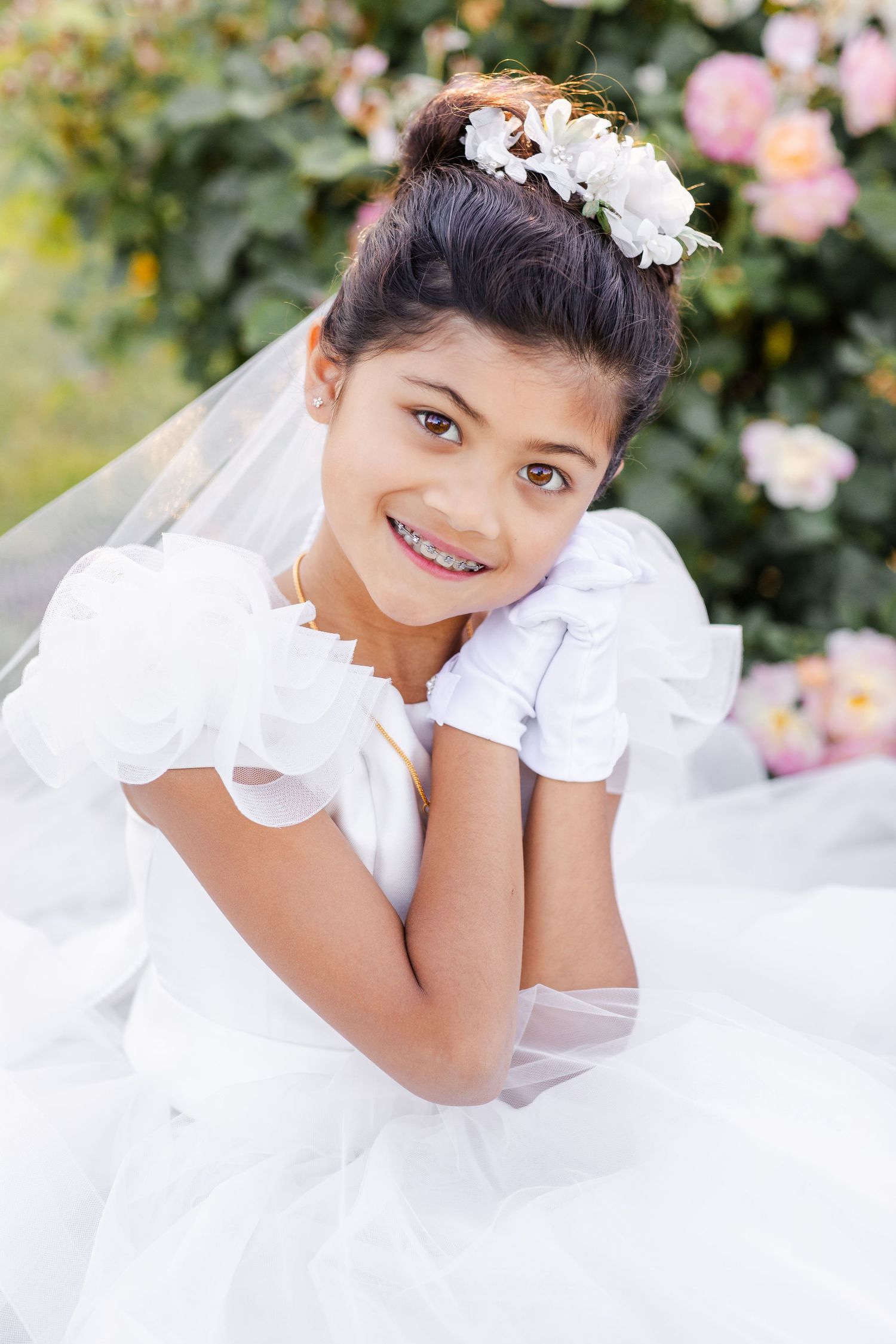 A young girl in a white First Communion dress and floral headpiece poses in front of blooming flowers.