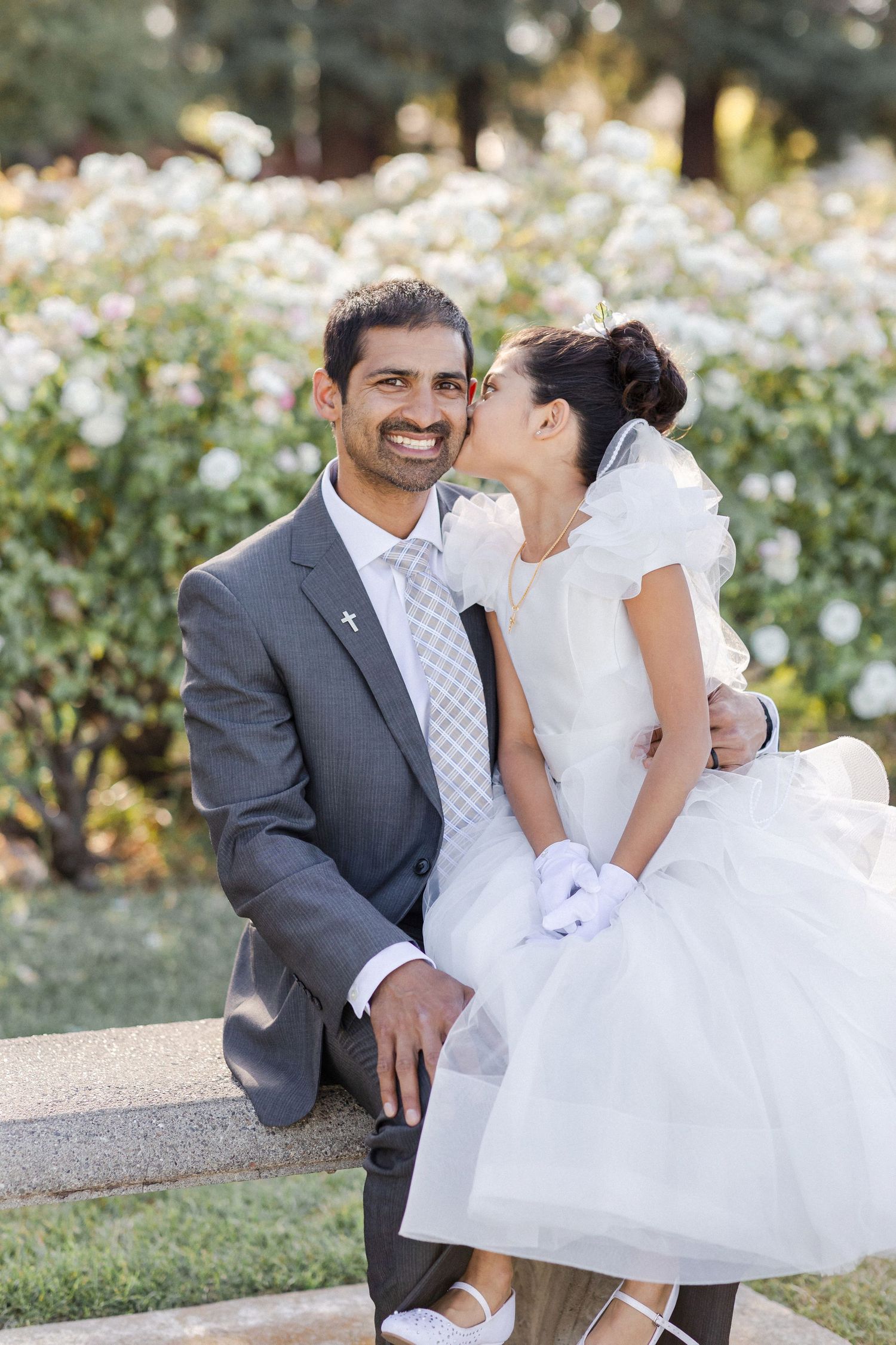 A couple in wedding attire share a tender moment together on a bench with white flowers blooming in the background.