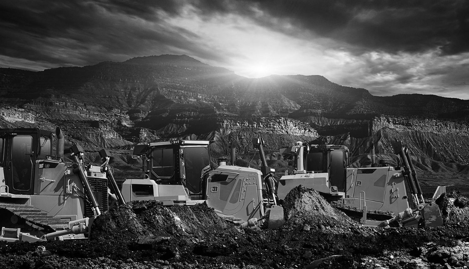 Black and white photo of heavy construction machinery at a mining site with dramatic mountain backdrop and sunset.