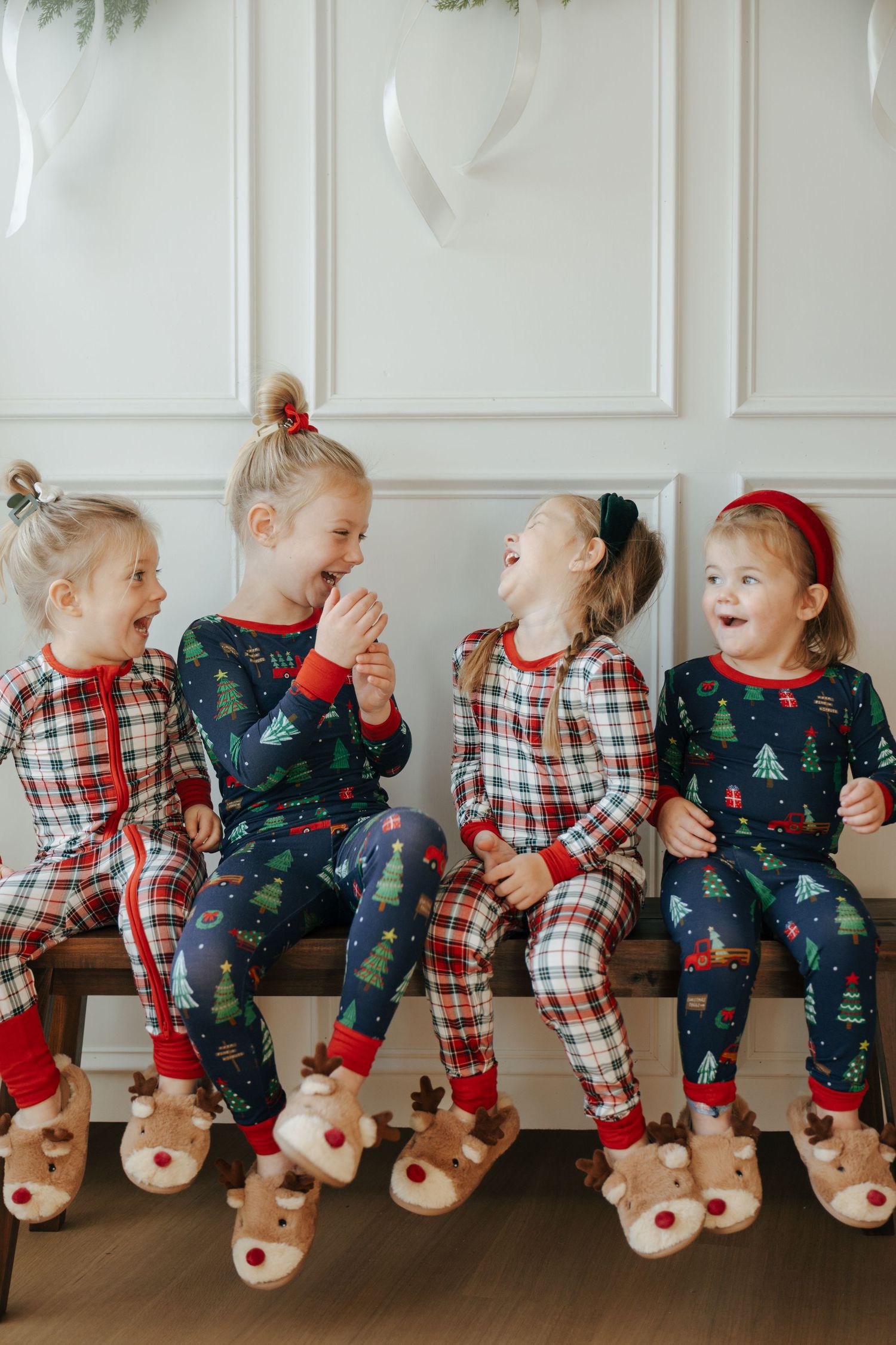 Children in matching Christmas pajamas sit in a row wearing reindeer slippers and laughing together.