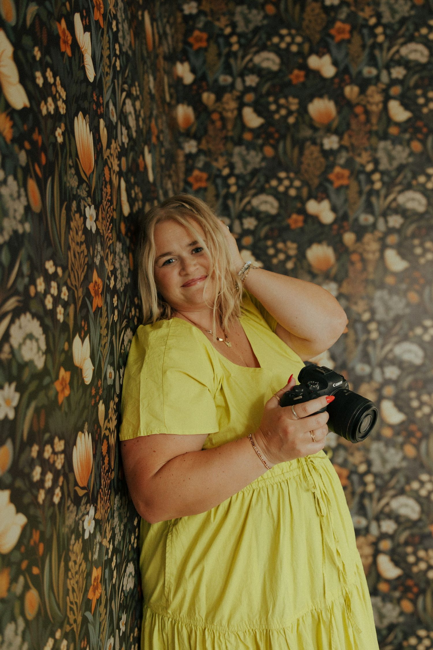 A photographer in a yellow dress poses with camera against floral wallpaper.