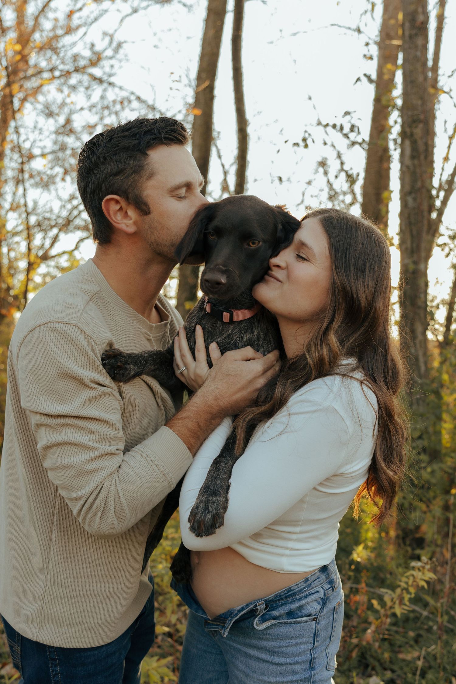 Couple sharing kisses with their black dog in autumn forest setting.