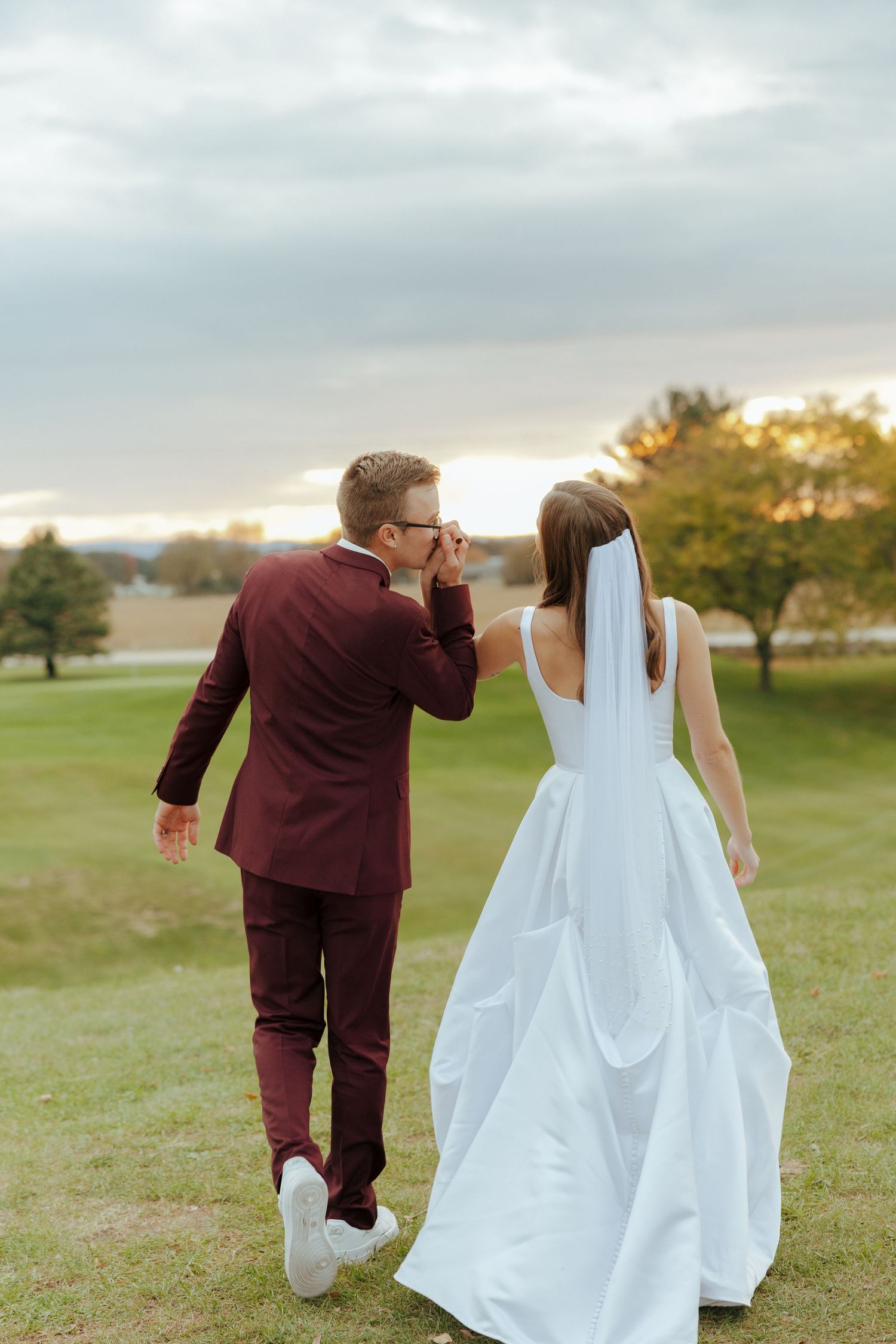 Bride and groom in burgundy suit running playfully across grassy field.