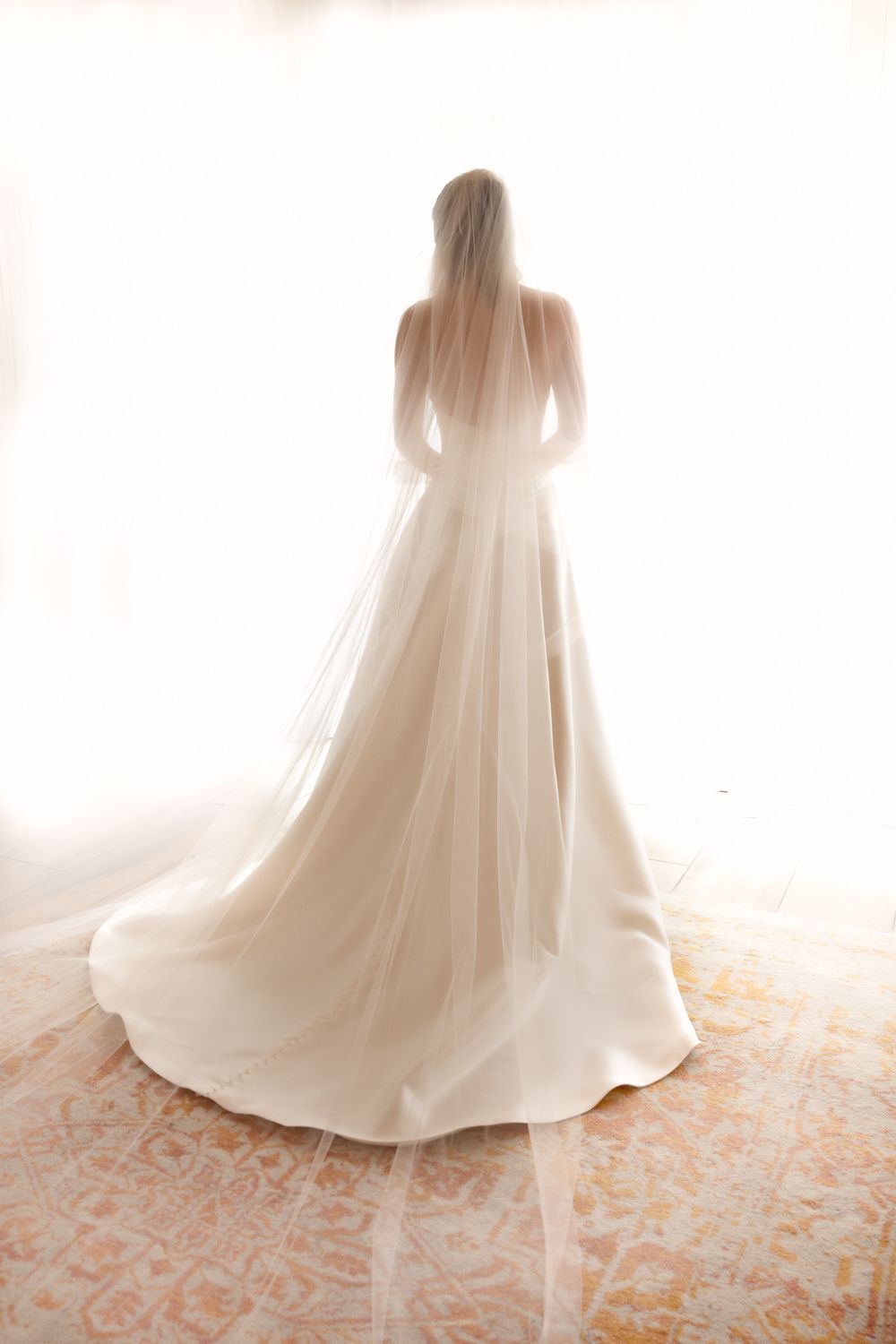 Bride in elegant white wedding gown with flowing veil and train, standing backlit near a bright window.