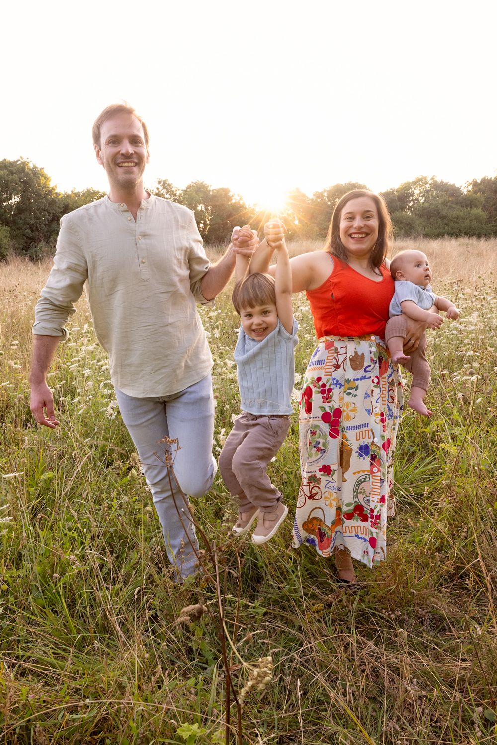 A family of four holding hands and walking through a sunny wildflower meadow at sunset.