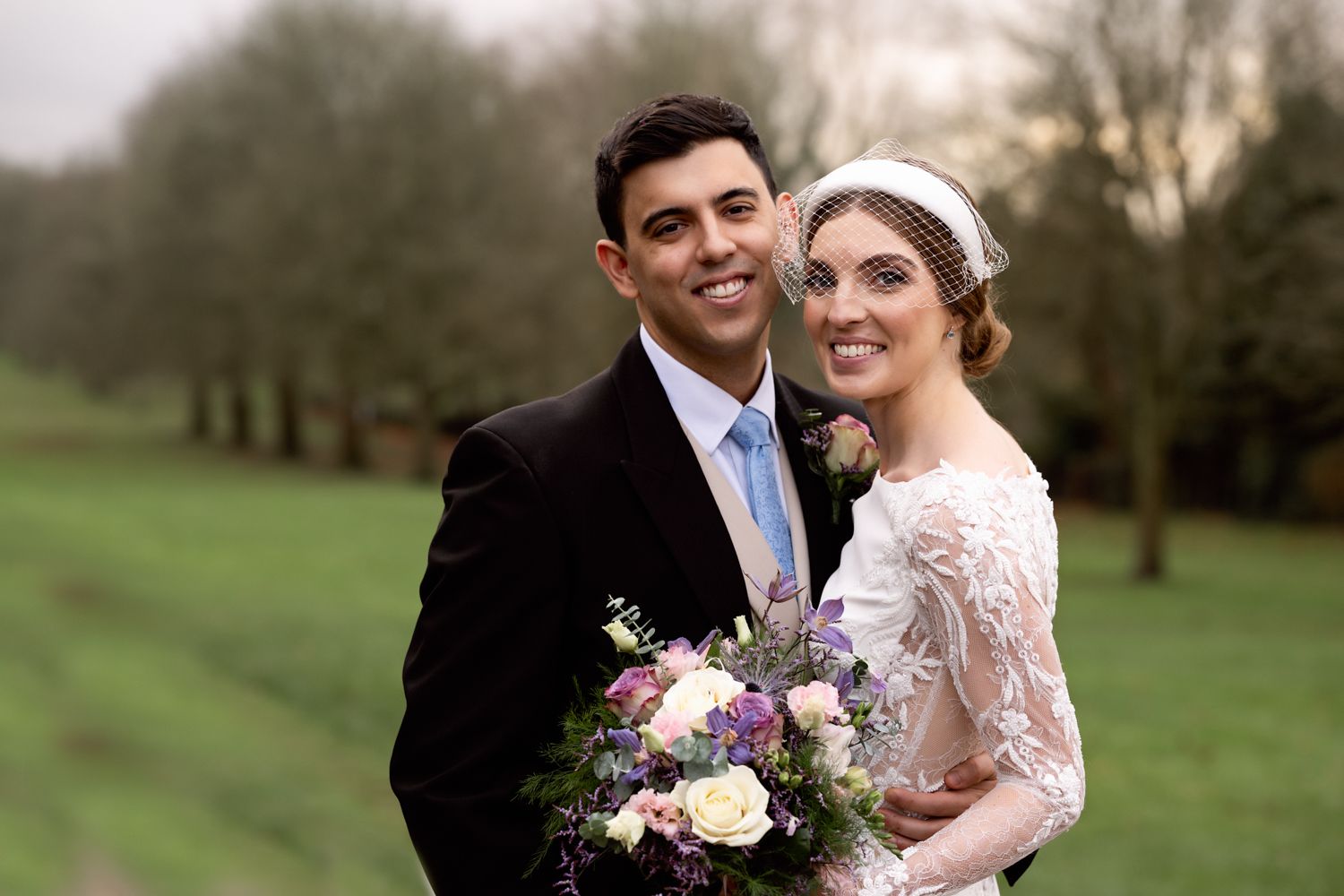 Newlywed couple poses outdoors holding a bouquet of pink and purple flowers against a dreamy nature backdrop.
