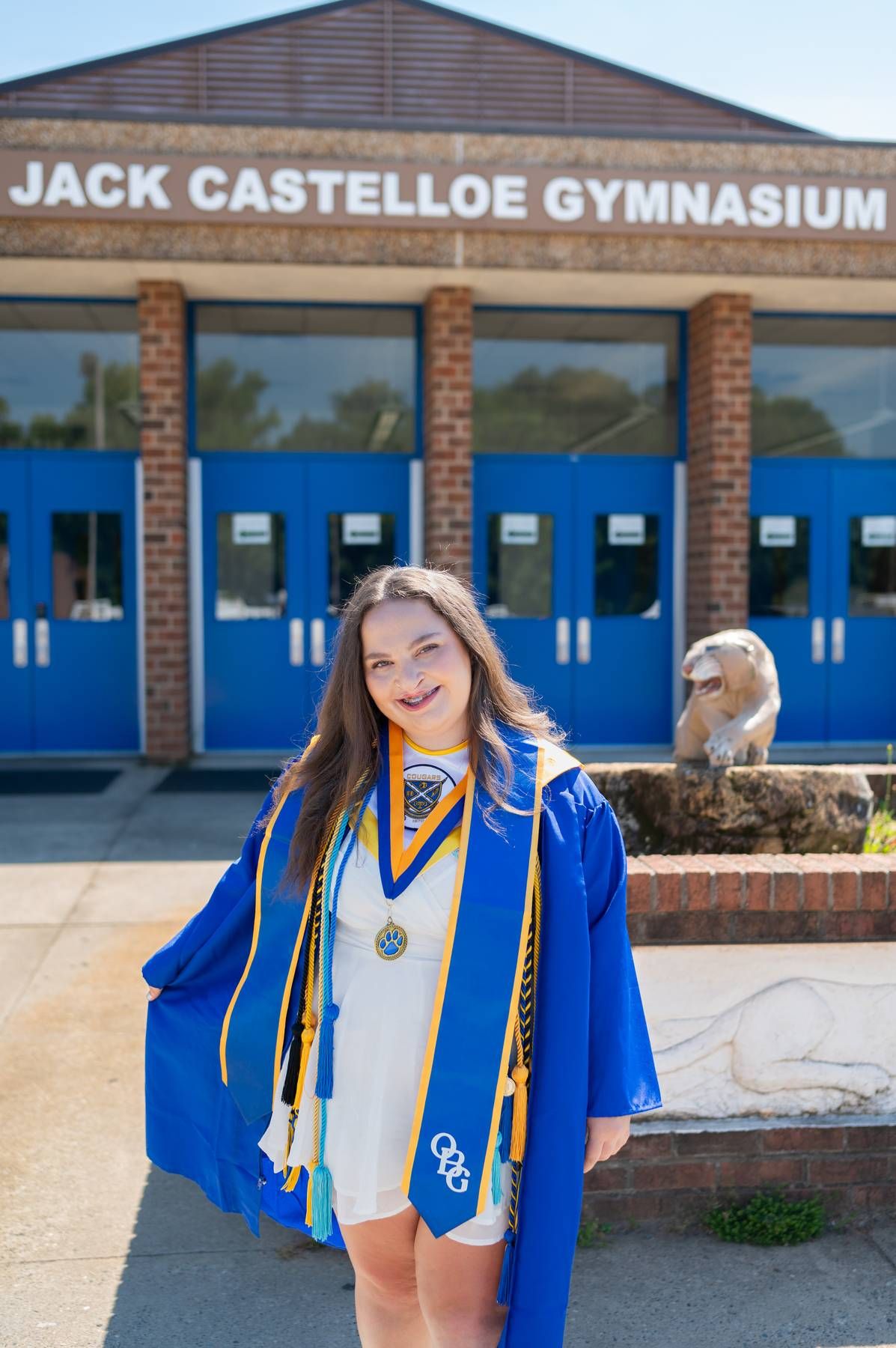 Smiling graduate in blue cap and gown stands outside Jack Castelloe Gymnasium, portrait photographer Asheboro NC