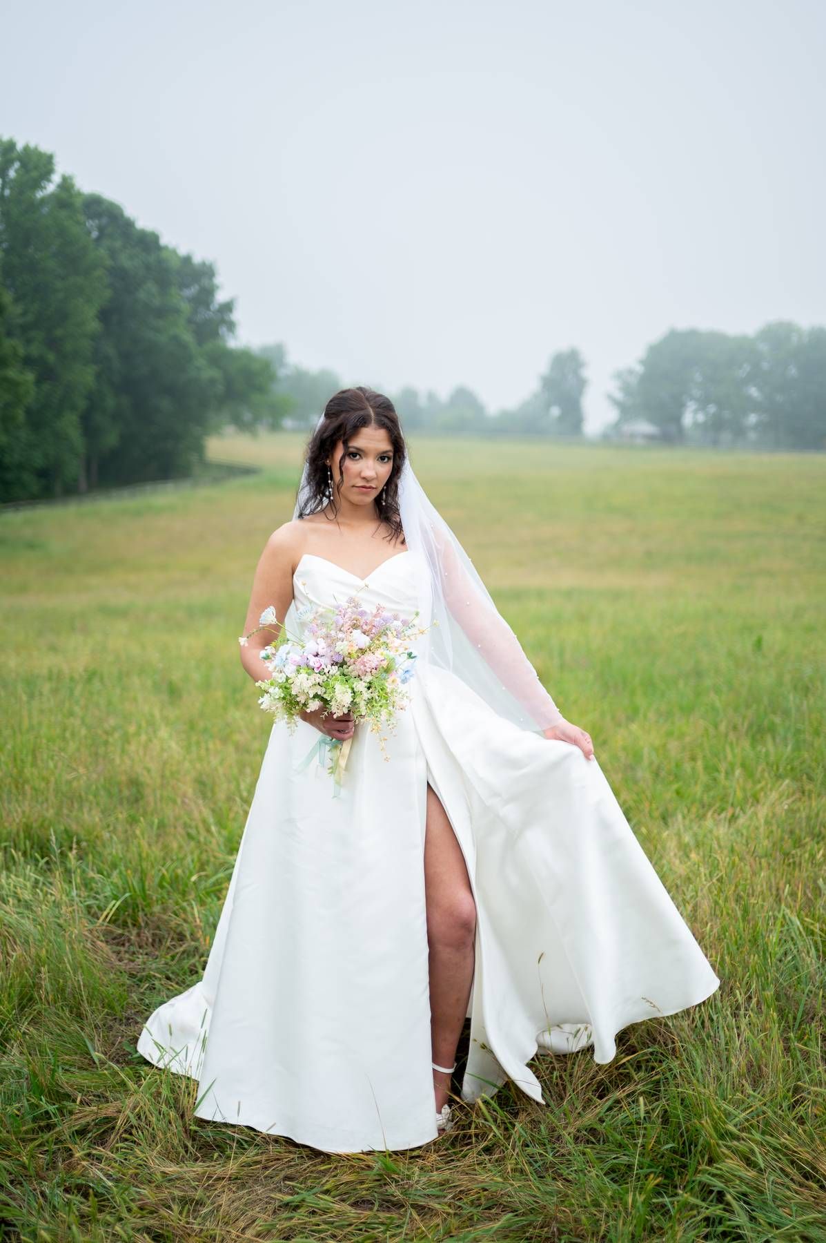 Bride in strapless white gown with slit holds a pink bouquet while posing in a misty green field with veil flowing, wedding photographer NC