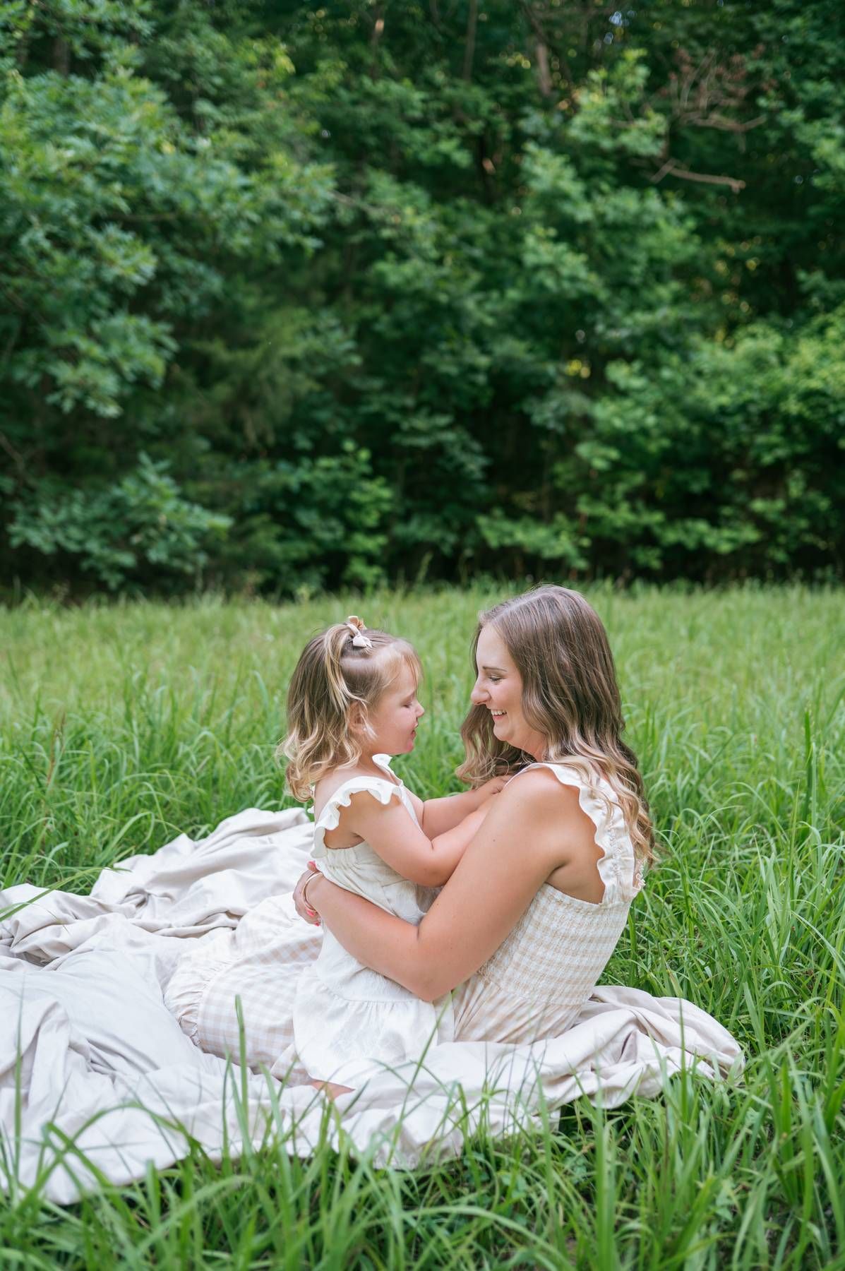 Mother and young daughter in white dresses sitting in tall green grass near trees, family photographer Lexington NC