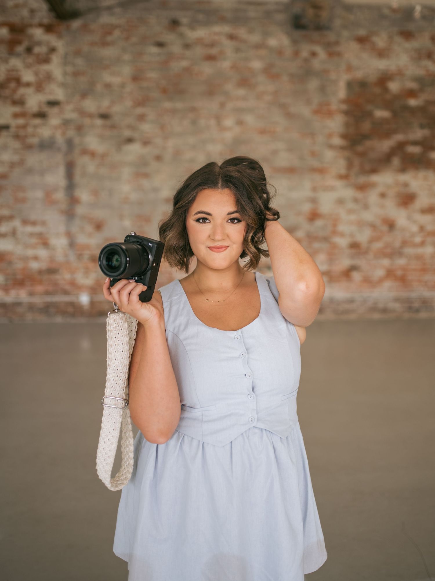 Smiling young woman in blue dress holds camera and woven bag in front of brick wall, wedding photographer NC