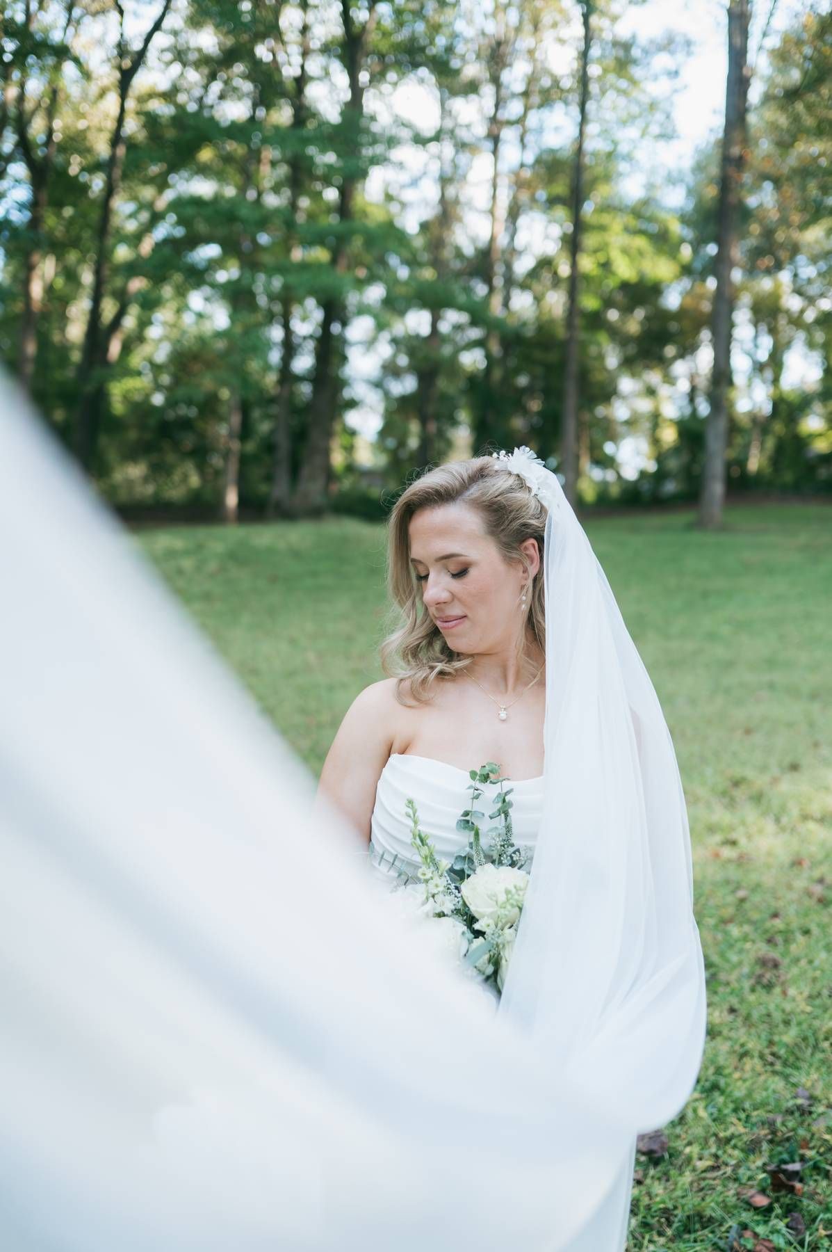 Blonde bride in strapless gown holding bouquet outdoors with veil flowing, wedding photographer High Point NC