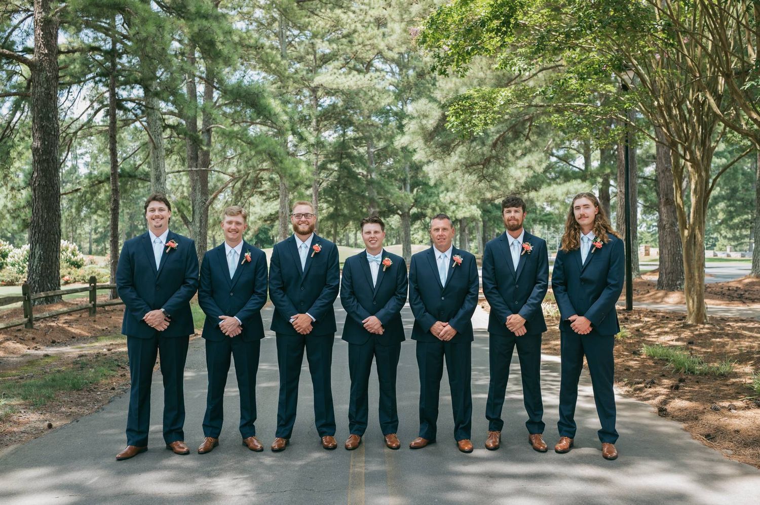 Seven groomsmen in navy suits posing together on a tree-lined path, wedding photographer Lexington NC